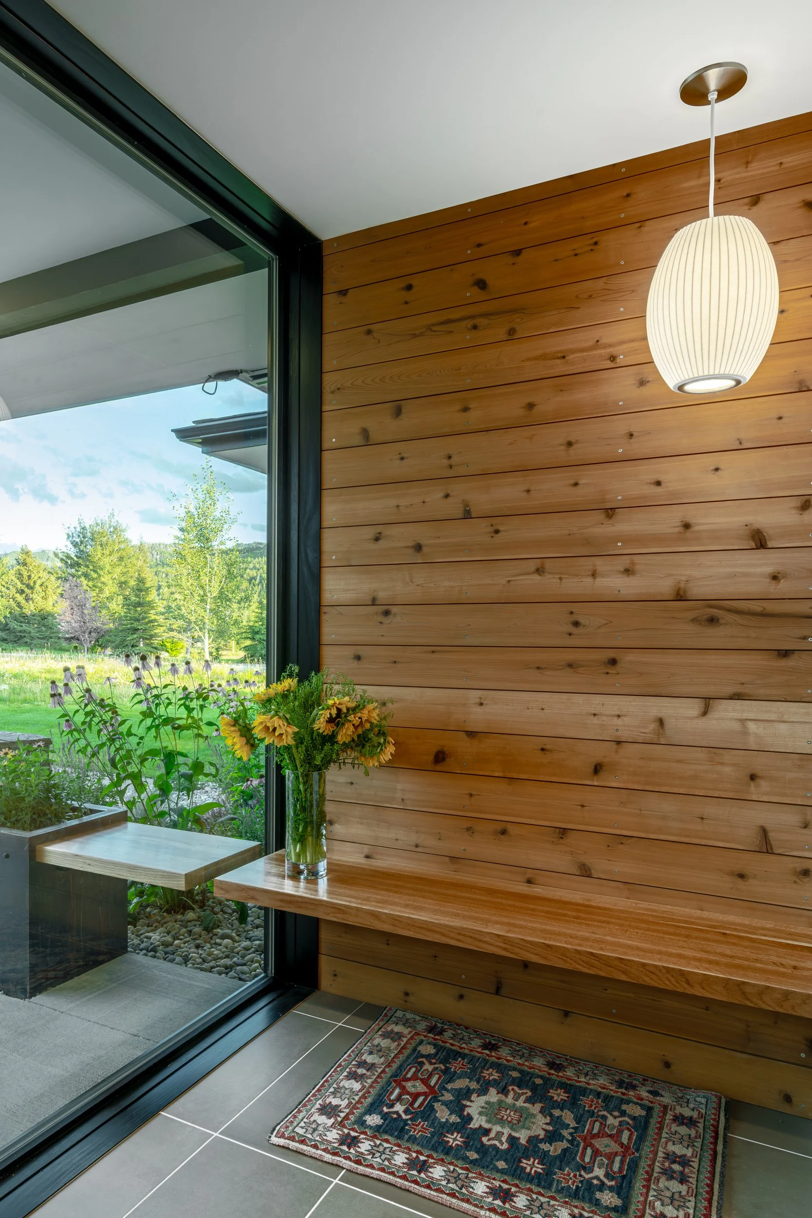Interior view of a room with a wooden wall, a hanging white paper lantern, a bench, a vase with sunflowers on a wooden table, tiled floor, and a sliding glass door to a garden with greenery and trees.