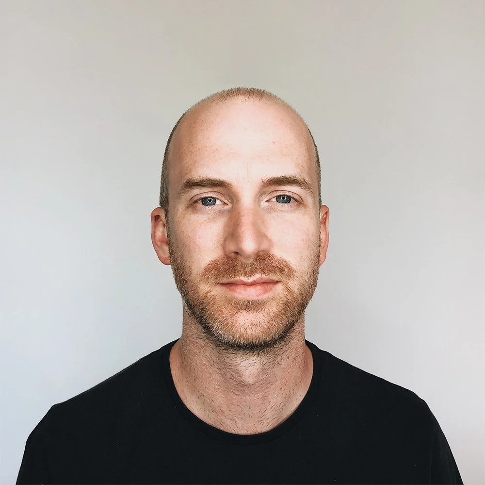 Portrait of a man with a light complexion, short light brown hair, blue eyes, and a beard, wearing a black shirt, against a plain light background.