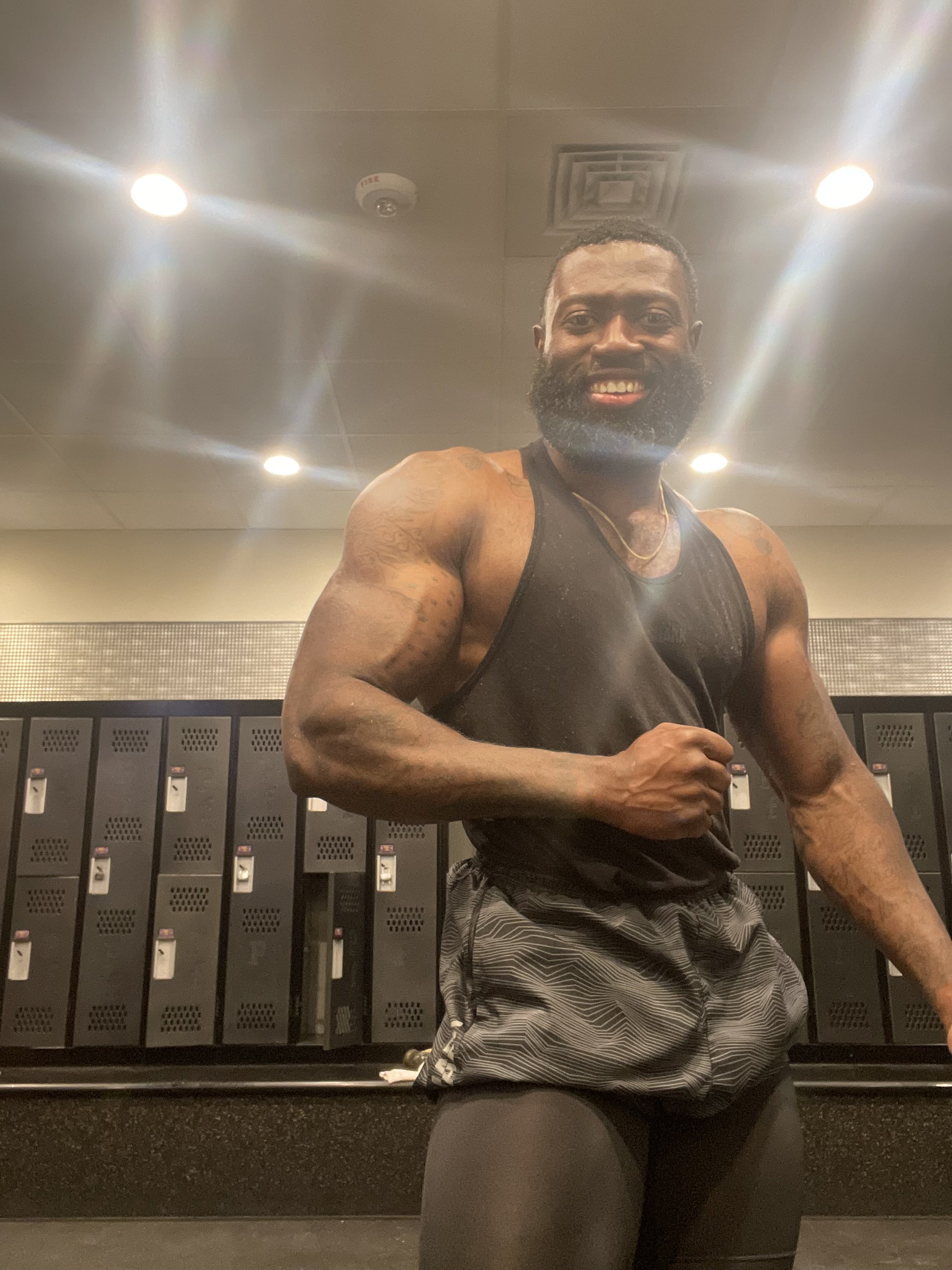 A muscular man with a beard in a gym locker room, smiling at the camera, wearing a black sleeveless athletic shirt and patterned shorts. Micah Sessoms