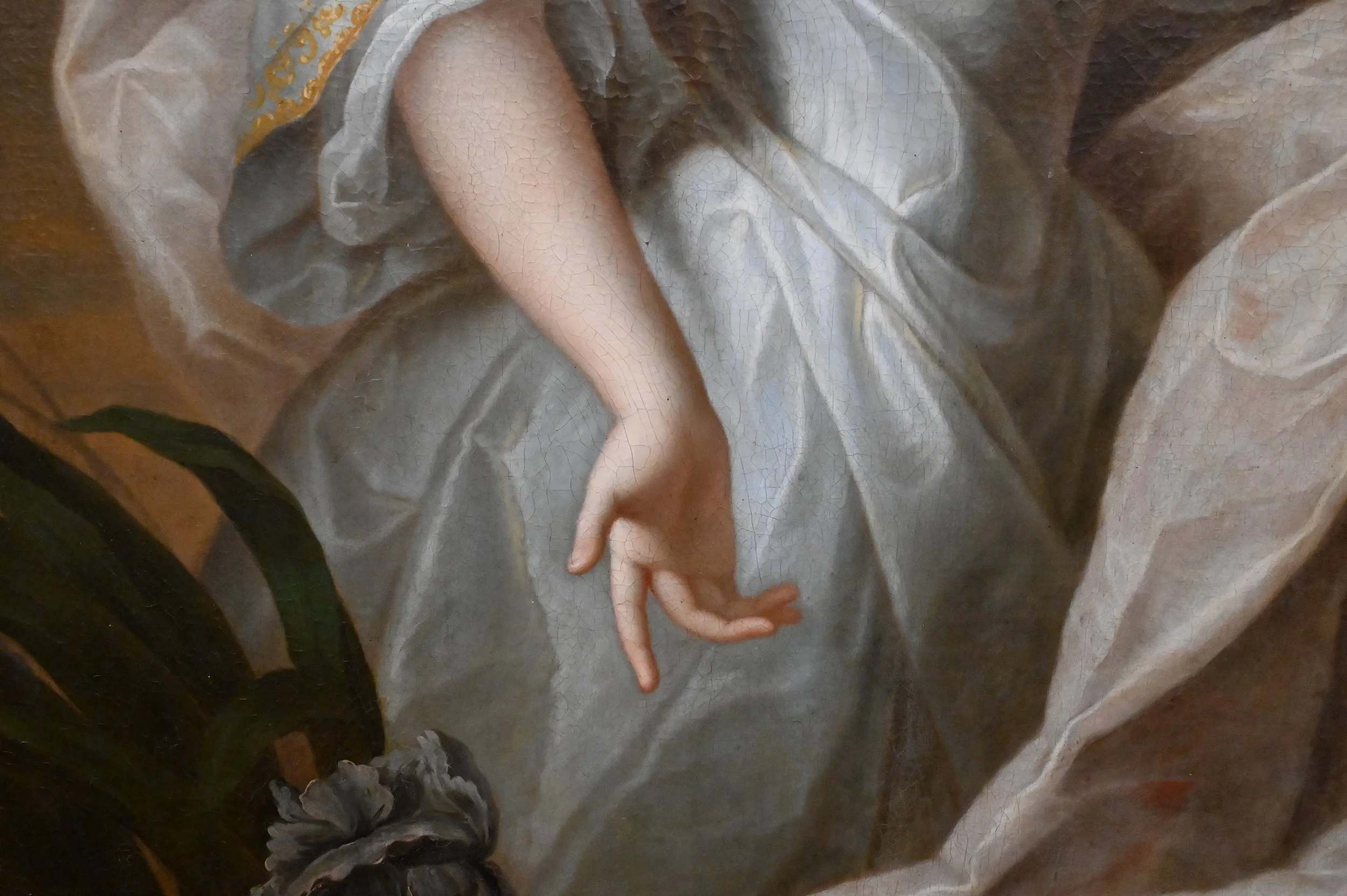 Close-up of a classical painting showing a hand with gentle fingers. The hand is part of a figure wearing flowing, light-colored fabric. There is a plant with dark green leaves and a flower at the bottom left corner.
