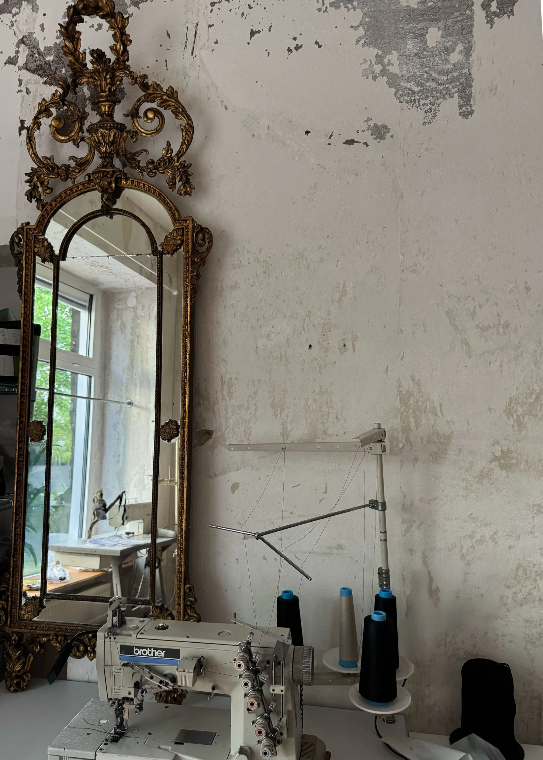 A sewing machine with large spools of thread, located in a room with an ornate gold-framed mirror reflection of a window, an unfinished wall, and various sewing supplies.