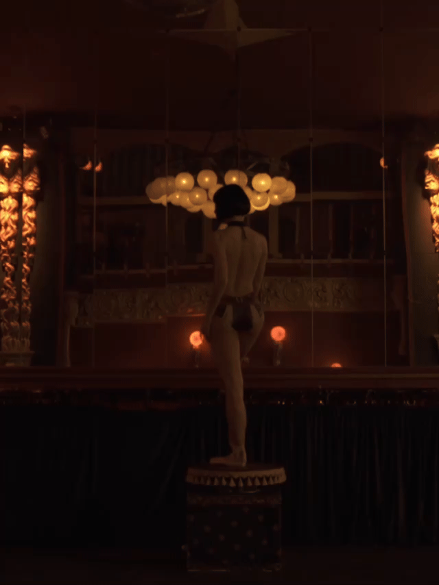 A woman standing on a pedestal in front of a mirror with ornate decor and warm lighting in a dimly lit room.