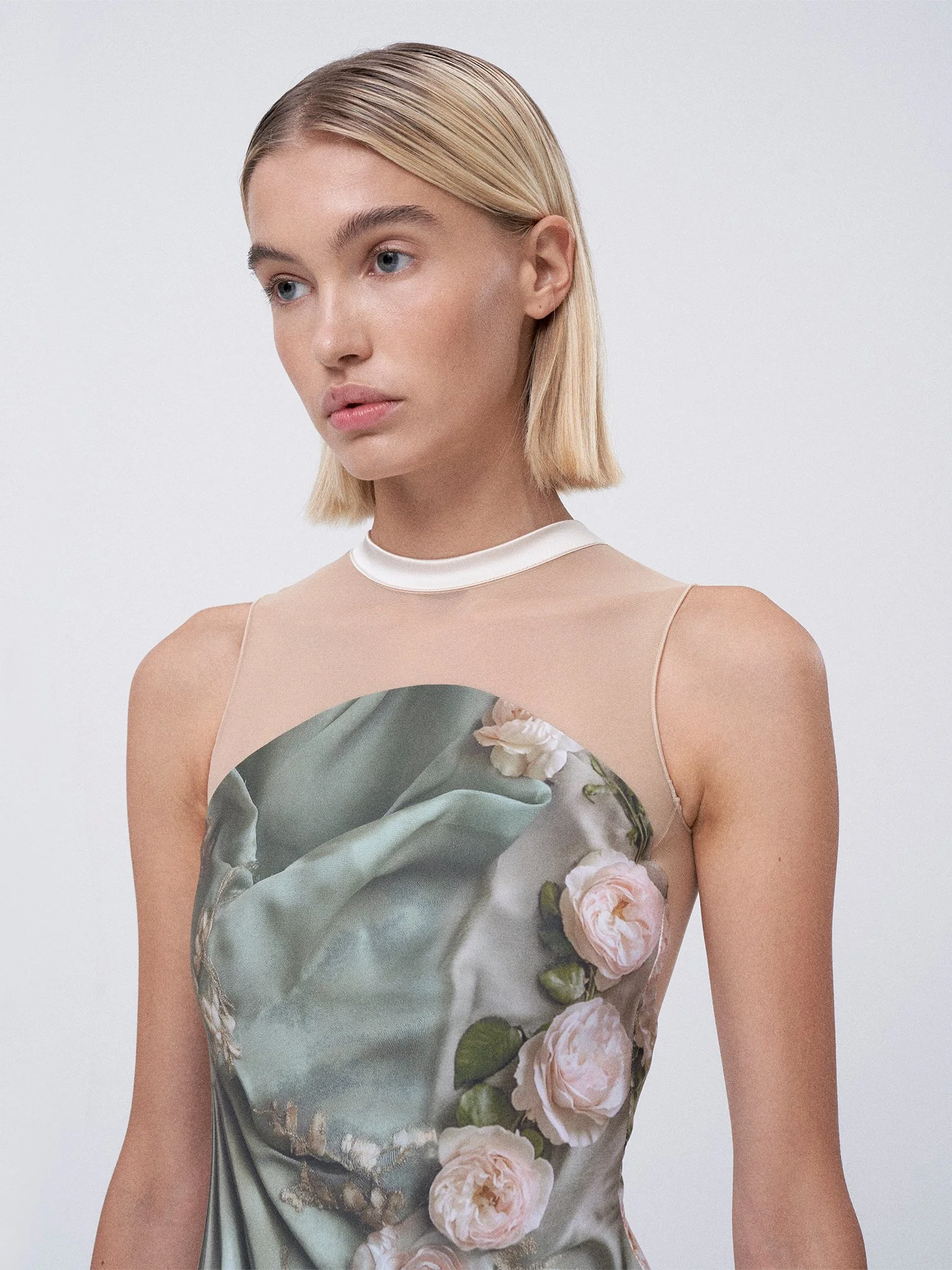 A young woman with blonde hair and blue eyes wearing a beige and floral dress with a mesh top and a pearl-like collar, gazing thoughtfully to the side against a plain white background.