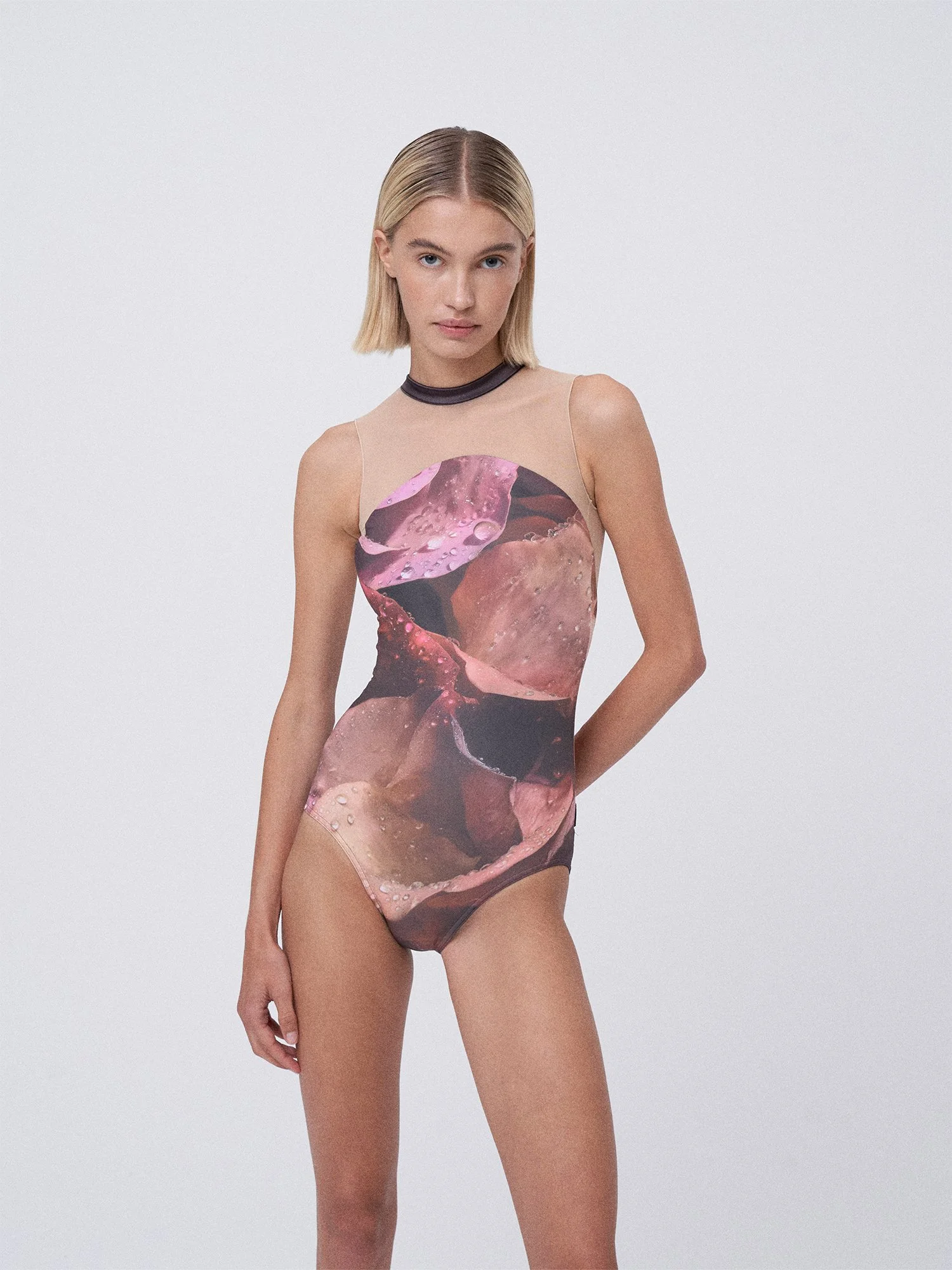 A young woman with blonde hair wearing a sheer, sleeveless, fitted dress with an abstract, pink and brown pattern, standing against a plain white background.