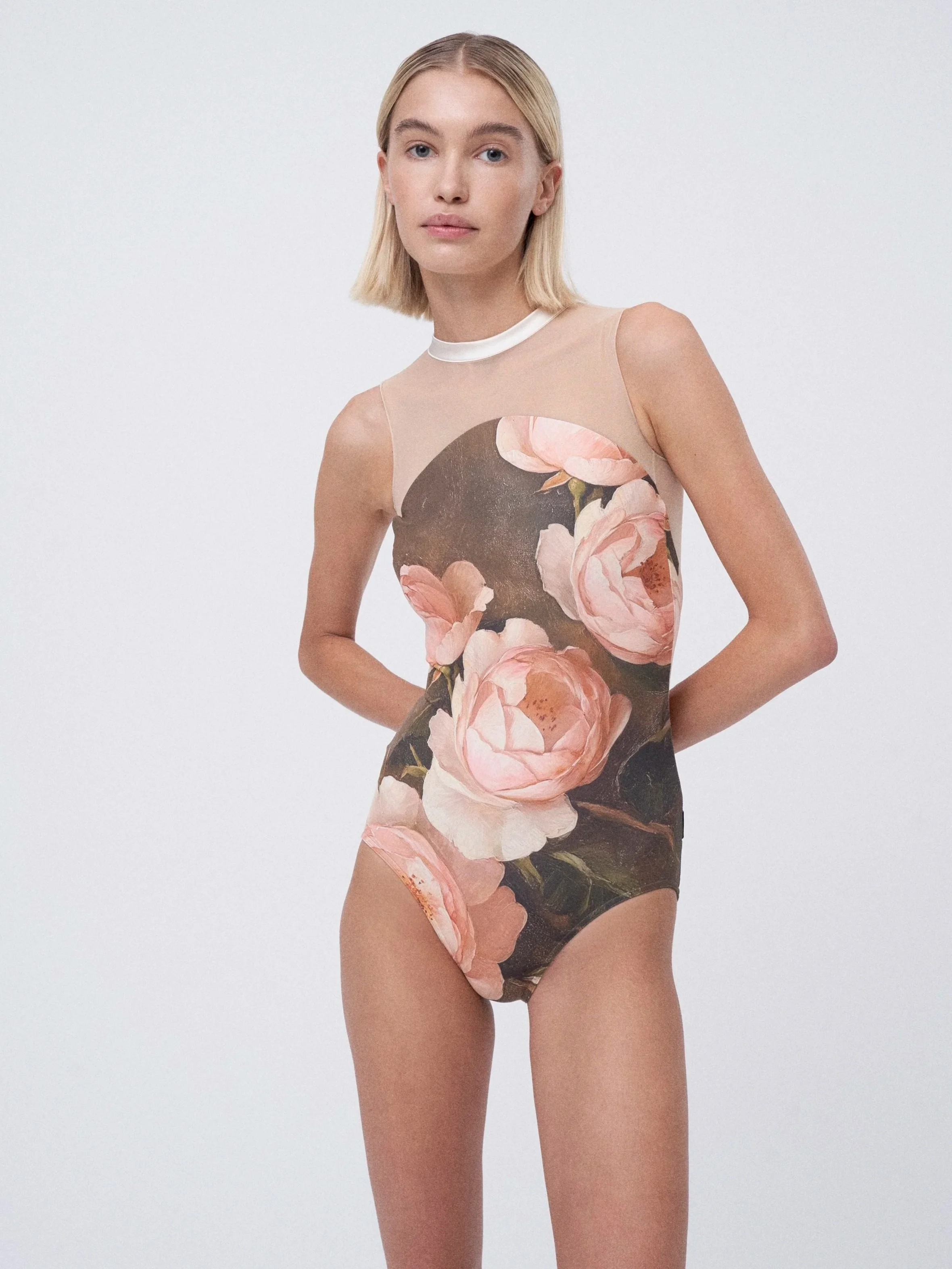 A young woman with blonde hair and a neutral expression wearing a sleeveless bodysuit with a large floral pattern of pink roses and dark leaves, standing against a plain white background.