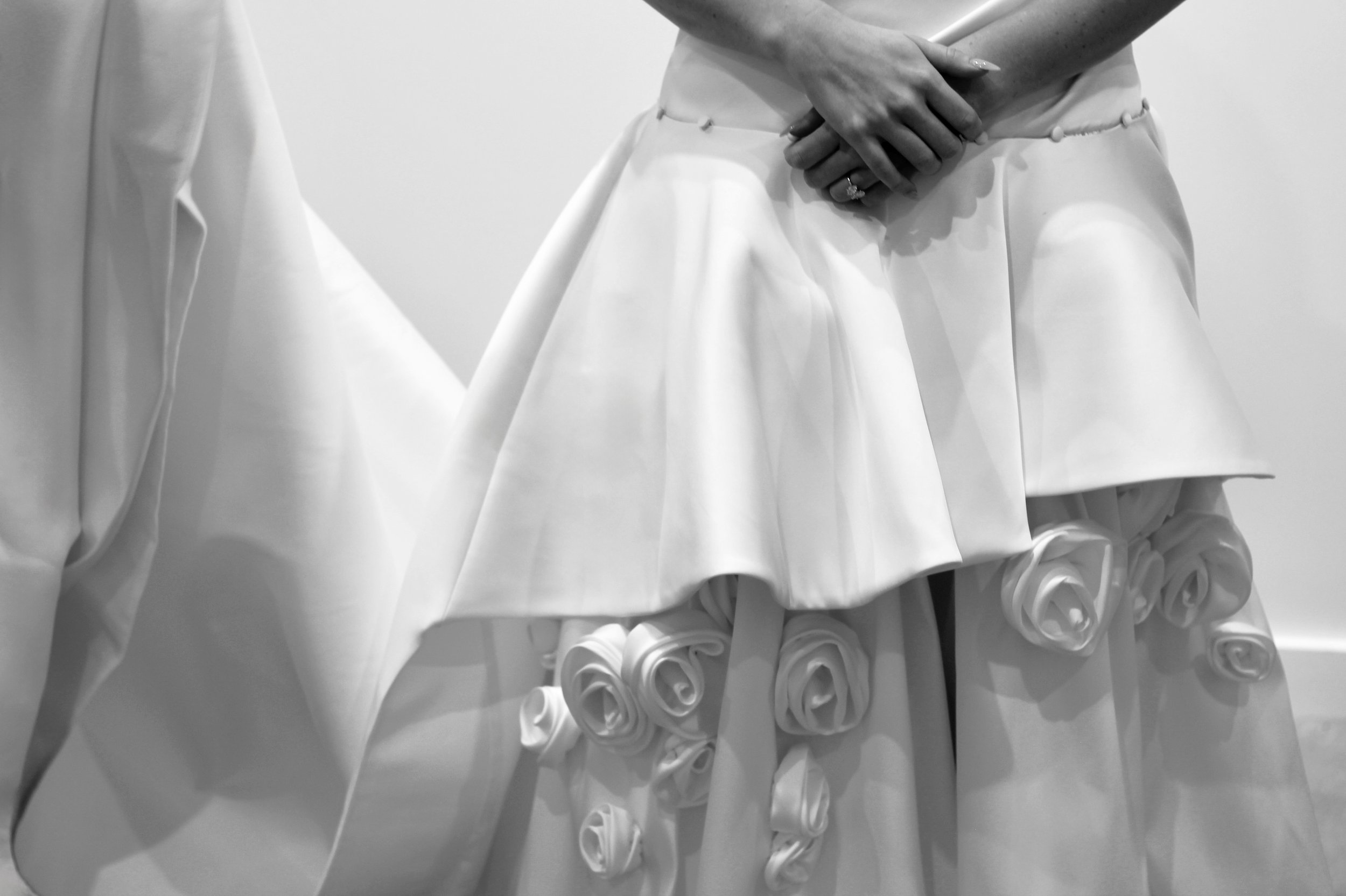 Close-up of a person wearing a white dress with multiple fabric roses at the bottom, hands clasped in front, rings visible.