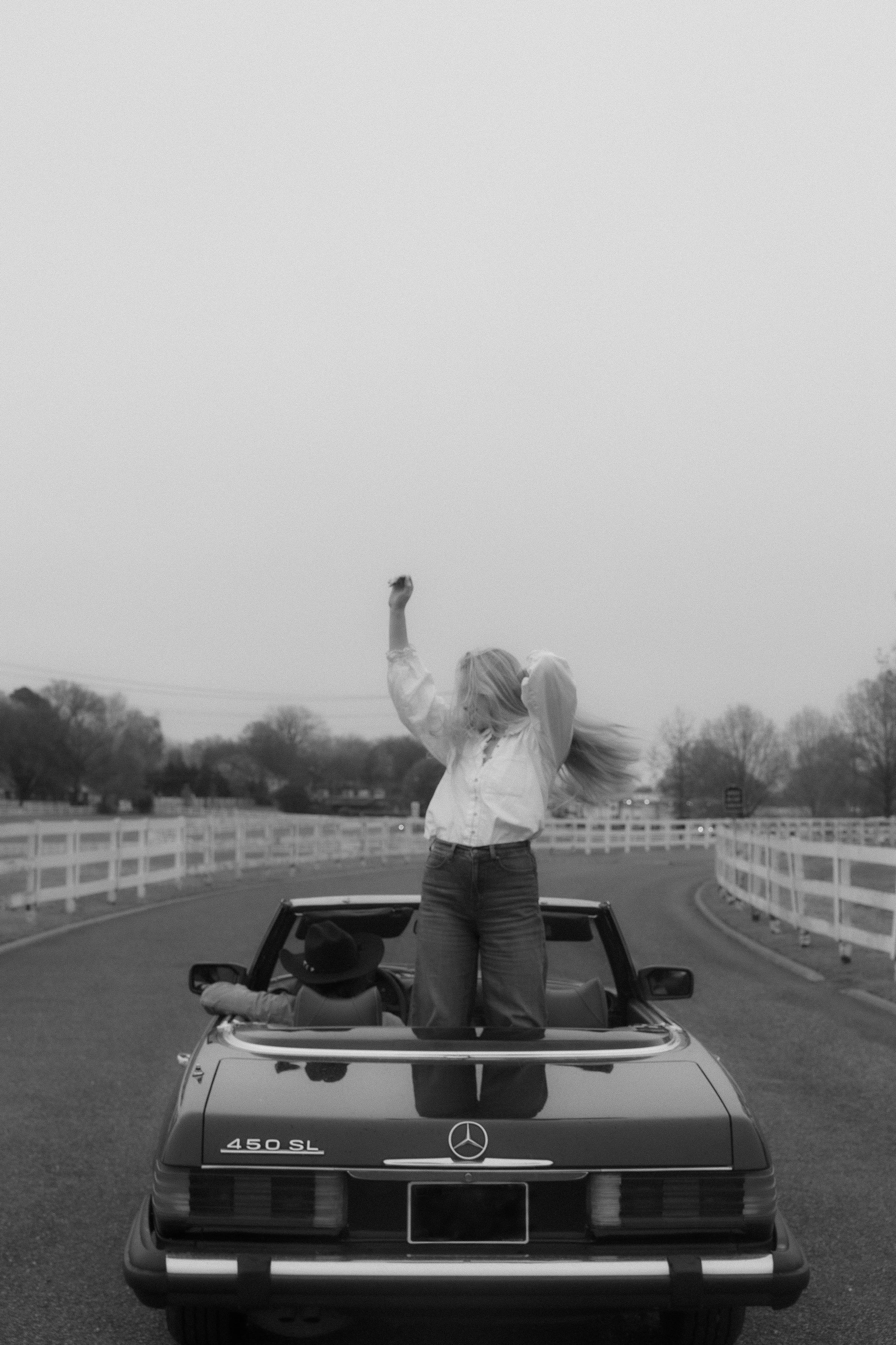 A woman standing on the back of a Mercedes-Benz 450SL convertible, raising her right arm in a fist, with her blonde hair flowing in the wind, as a person wearing a cowboy hat is sitting in the driver's seat. The scene is on an empty curving road with white fences on both sides, and trees in the background.