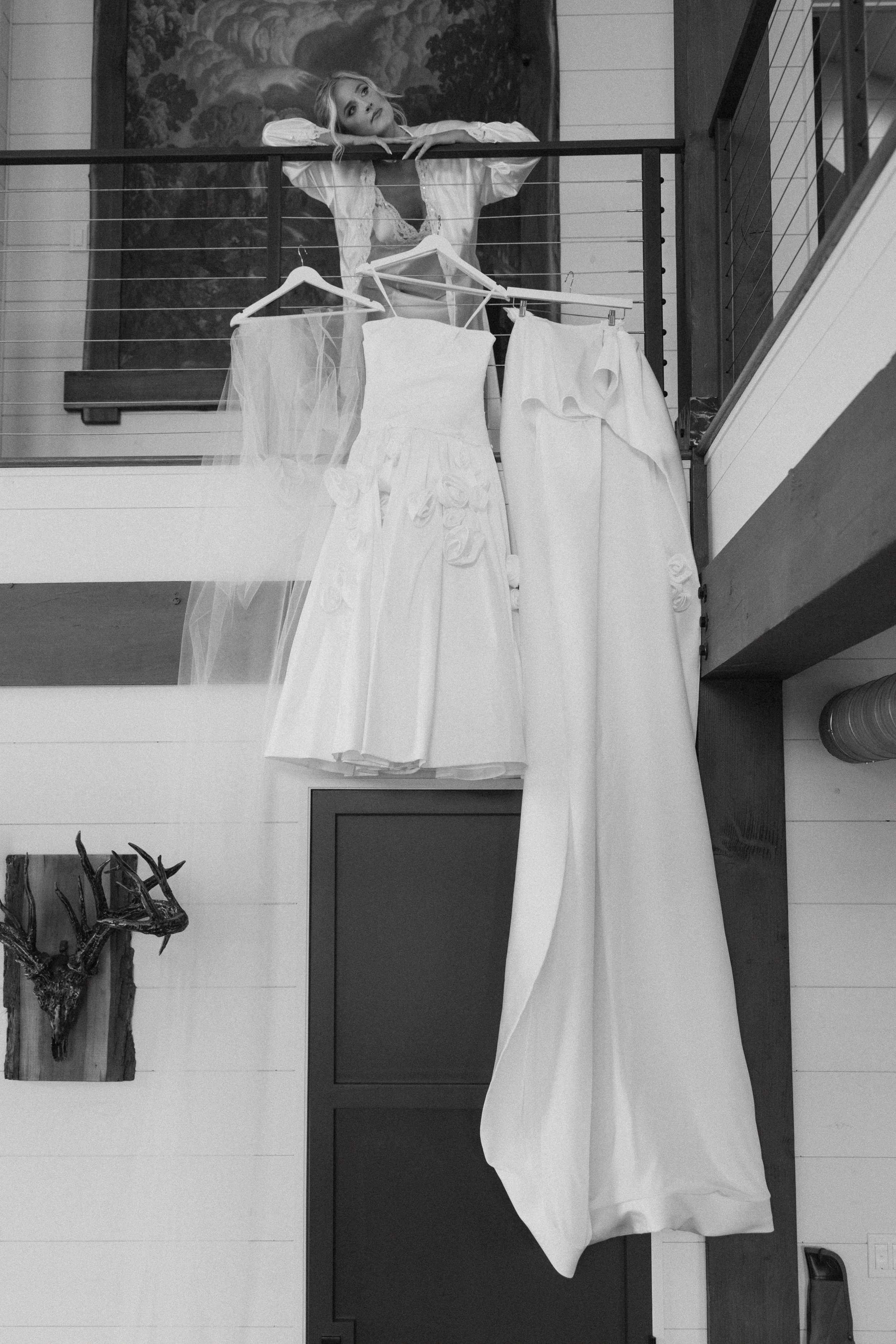 Black and white photo of a woman leaning on a balcony railing, looking to the side, with three white dresses hanging in front of her.