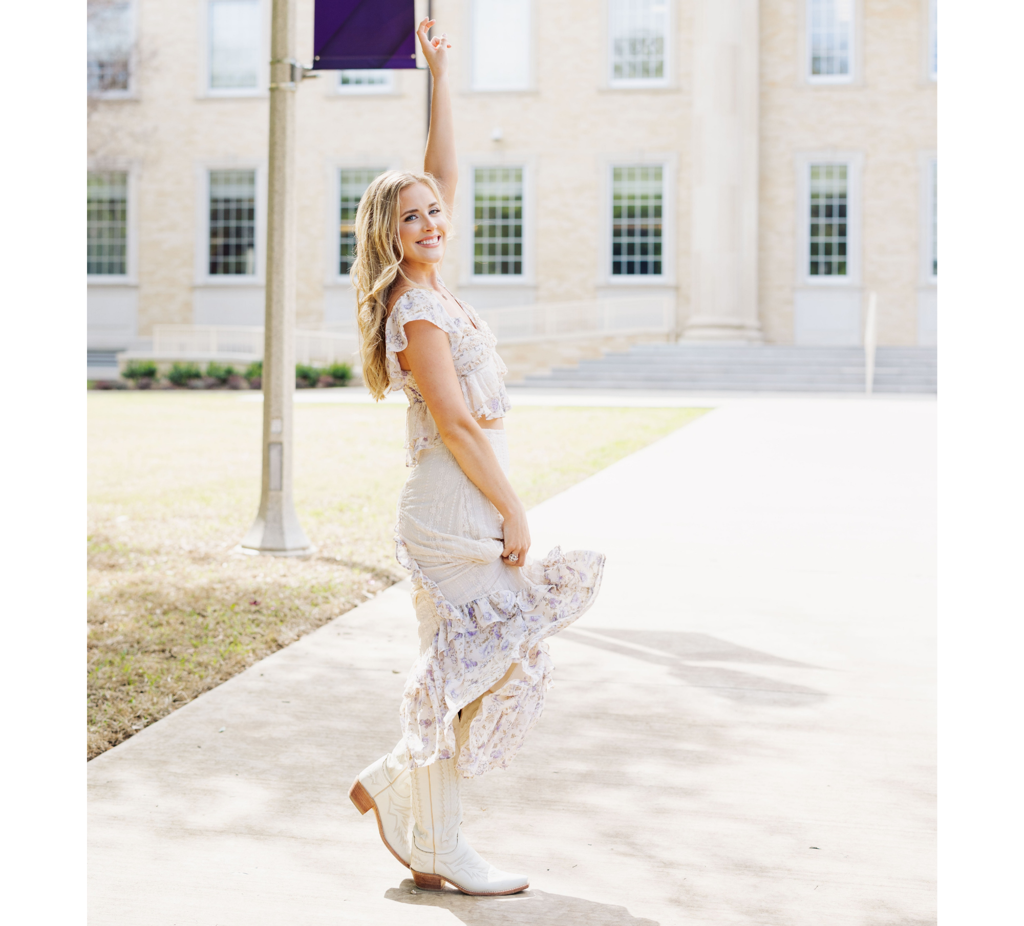 A young woman with long blonde hair smiling and twirling outdoors near a large building, wearing a flowy floral dress and white cowboy boots.