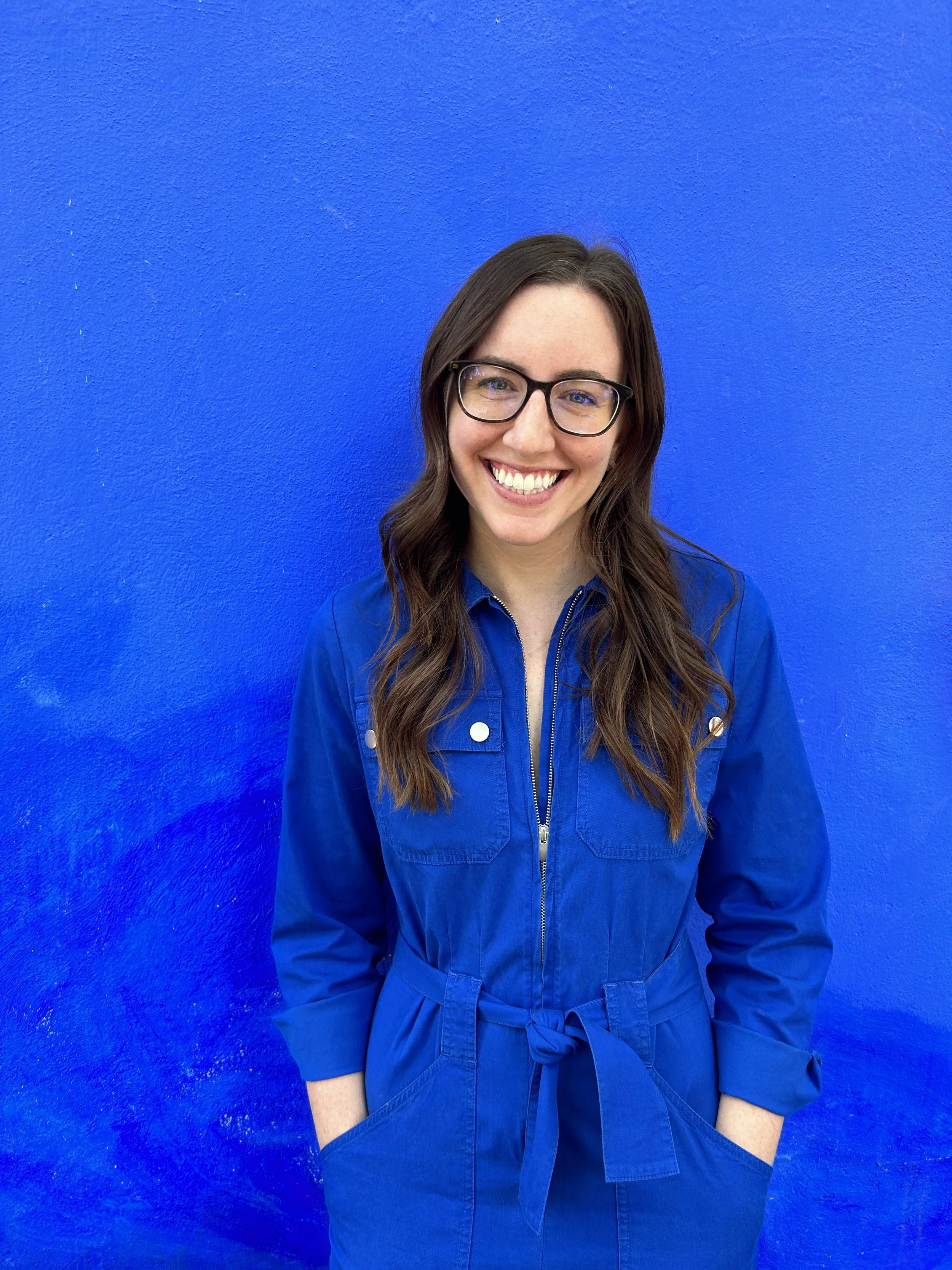 A woman with long wavy brown hair, wearing glasses and a blue jumpsuit, smiling and standing in front of a solid blue wall.