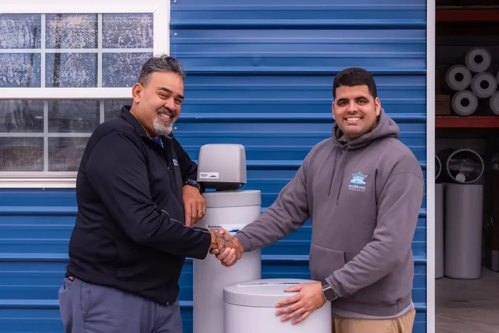 two men smiling, one wearing a grey hoodie with rainking logo on the right and one with a black hoodie on the left, shaking hands over water softener and salt tank. backgorund is a home with blue metal panneling