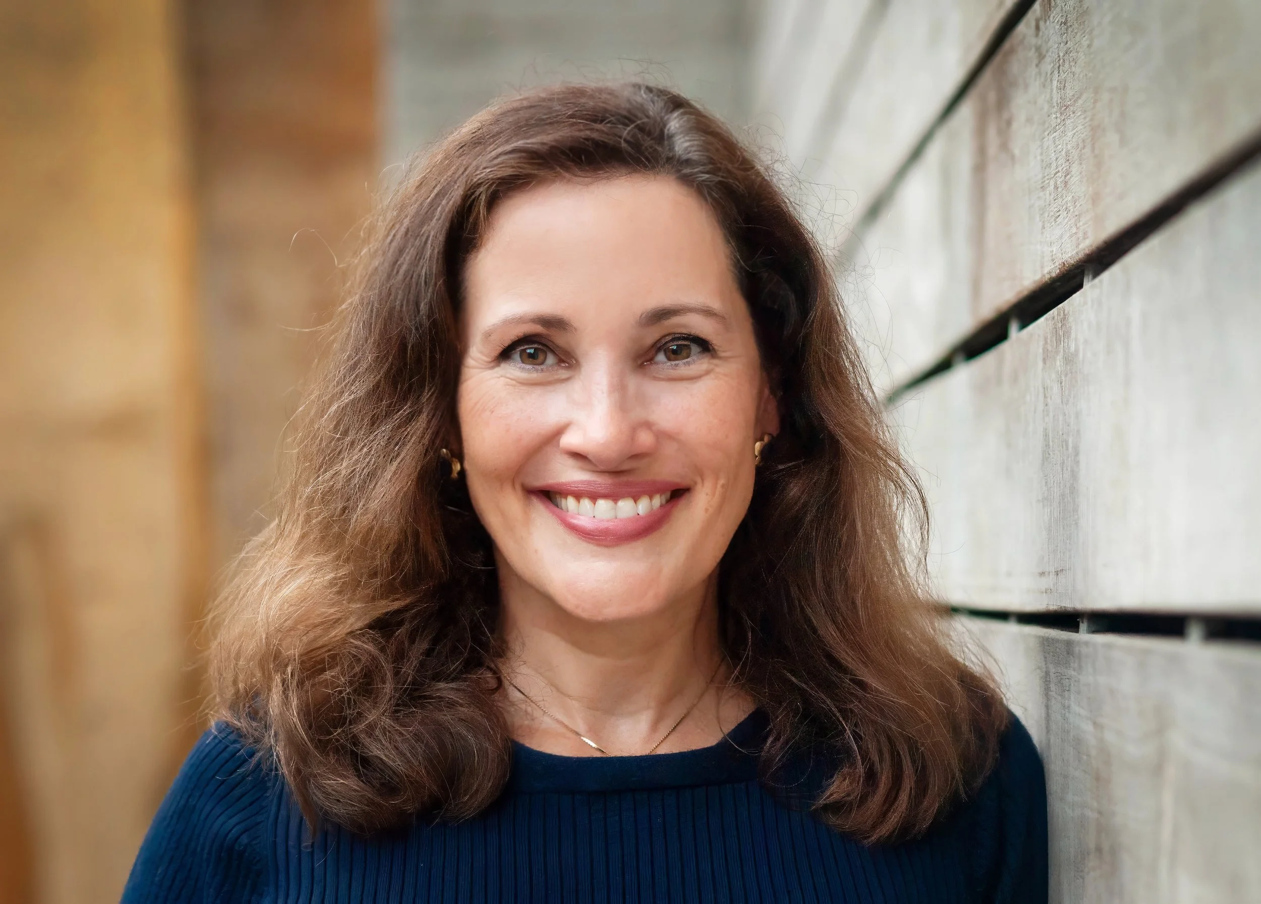 Close-up of a smiling woman with brown curly hair, wearing a navy blue top, standing against a wooden wall.