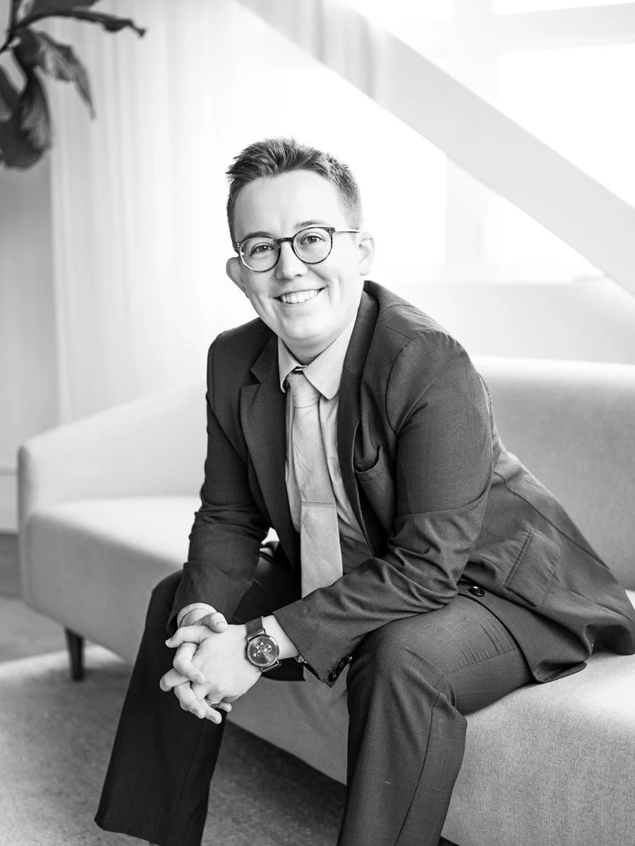 A smiling person with short hair and glasses, dressed in a suit and tie, sitting on a couch in a bright, modern room.