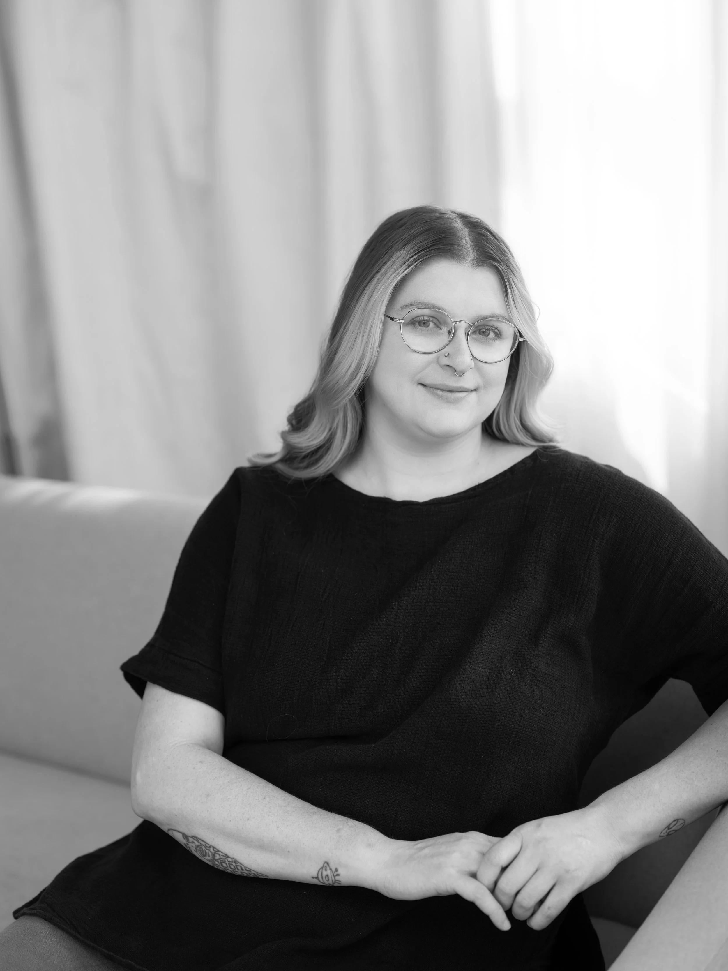 Black and white photo of a young woman with glasses, wavy hair, and tattoos on her arms, sitting with her arms crossed in front of her, smiling slightly, in a well-lit room with curtains in the background.