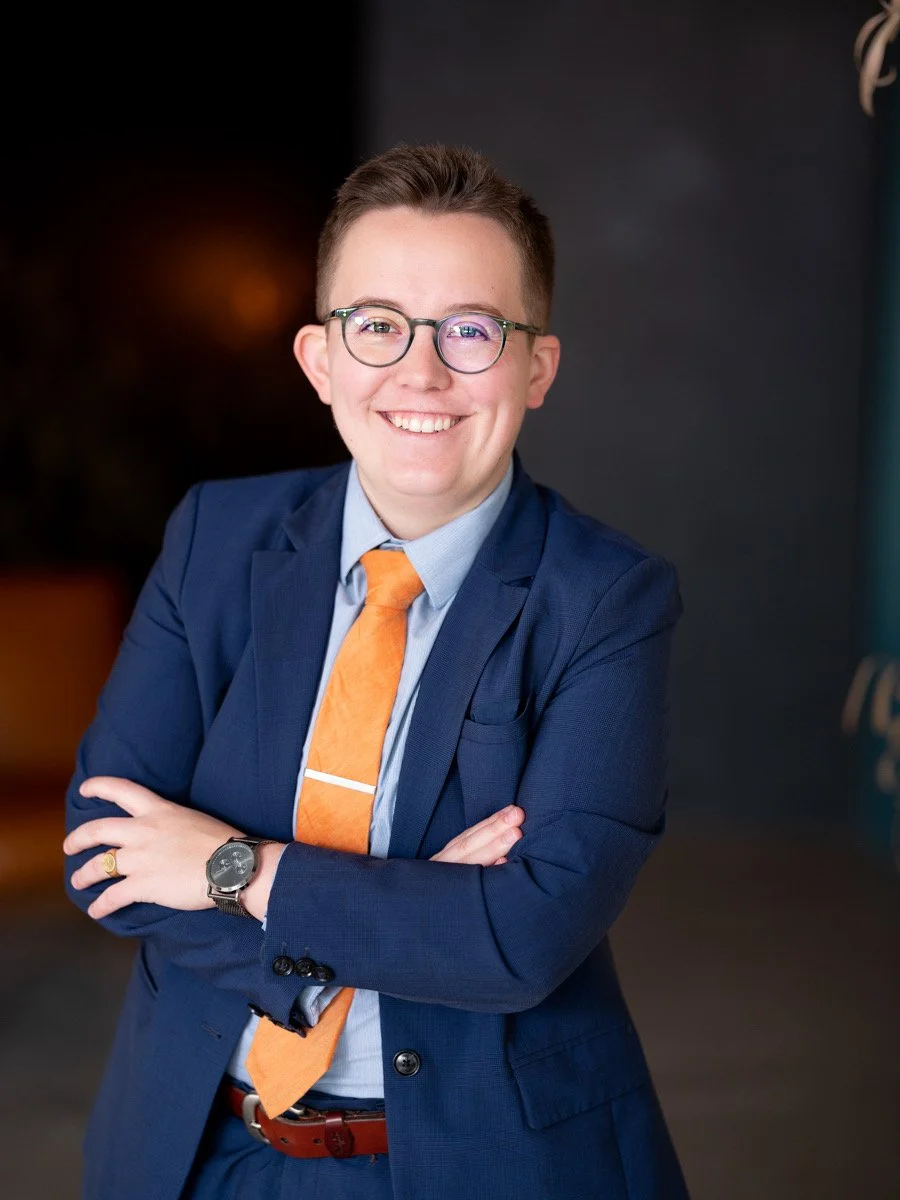 A smiling woman with short brown hair and glasses, wearing a blue suit, light blue shirt, and orange tie, posing with her arms crossed in a professional setting.