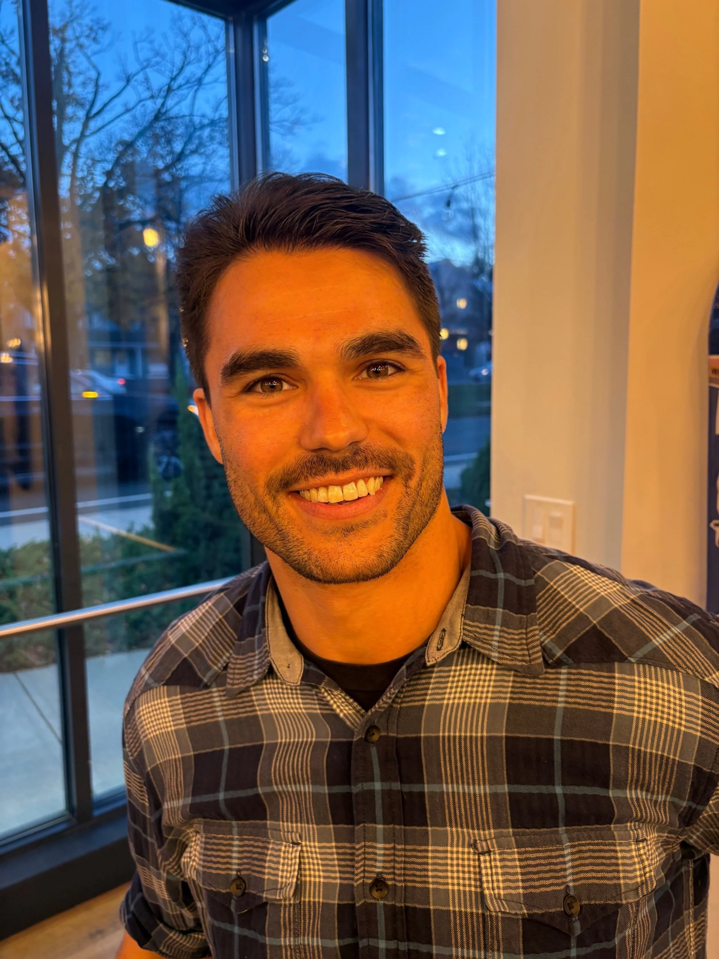 A smiling man with dark hair and a beard wearing a plaid shirt inside a room with large window showing trees and houses outside at dusk.