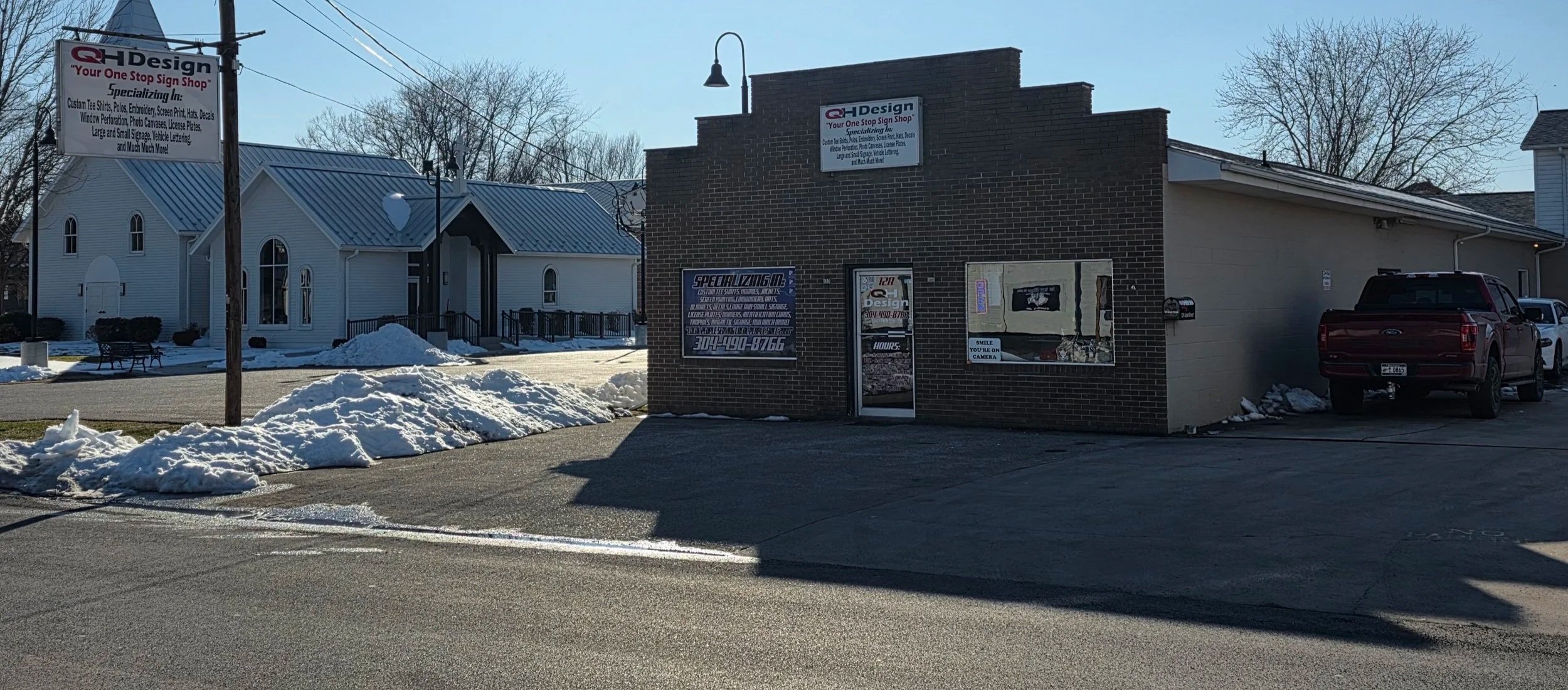 A small commercial building with signs for sign design and specialty services, and a parking lot with a red pickup truck. Snow piles are along the sidewalk, and a church with a metal roof is in the background.