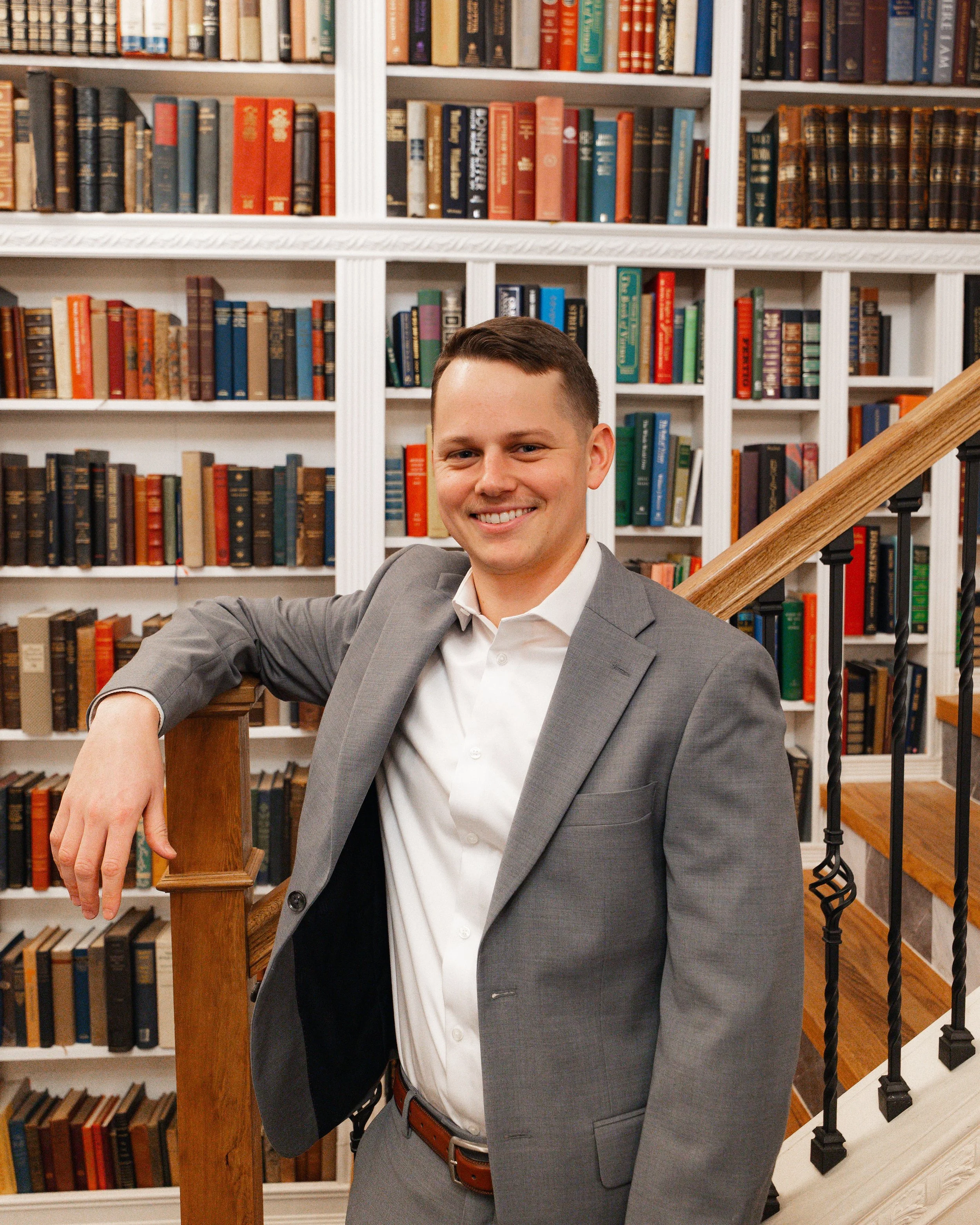 A young man in a gray suit and white shirt smiling in a library with shelves of books in the background.