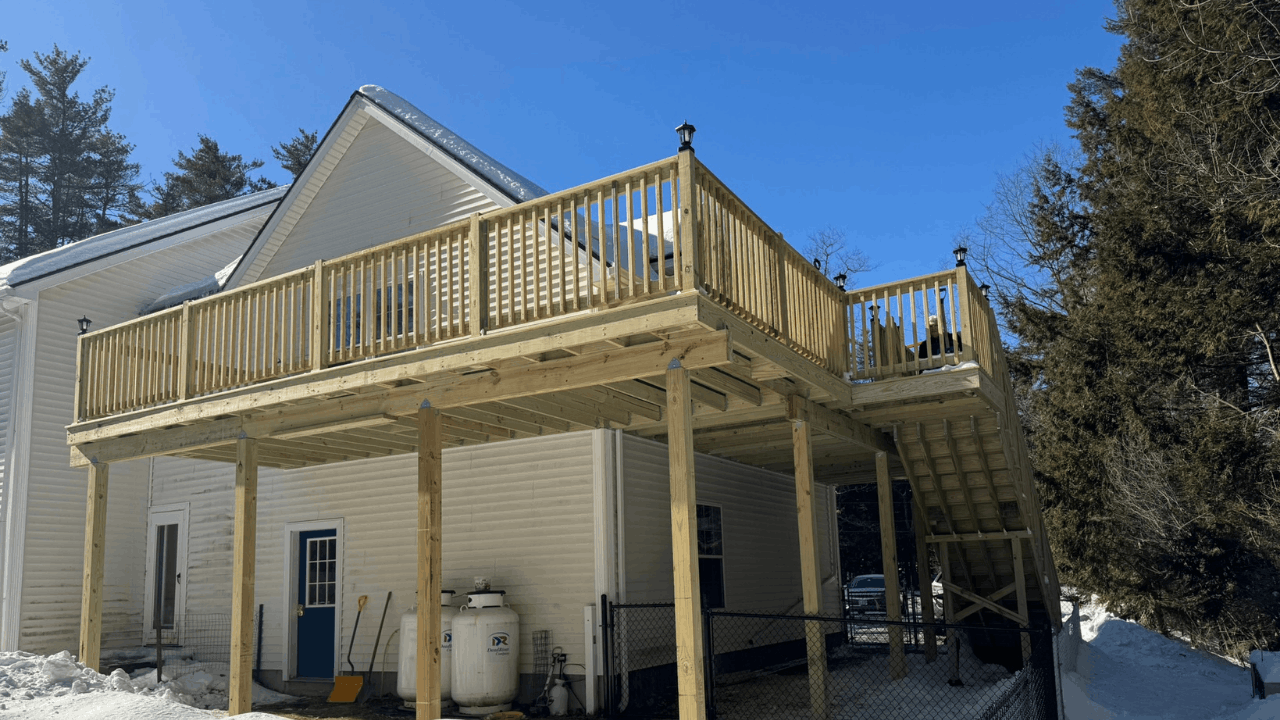 Newly built wooden deck on the second floor of a house, with stairs leading down to the ground, winter snow on the ground, and surrounded by trees.