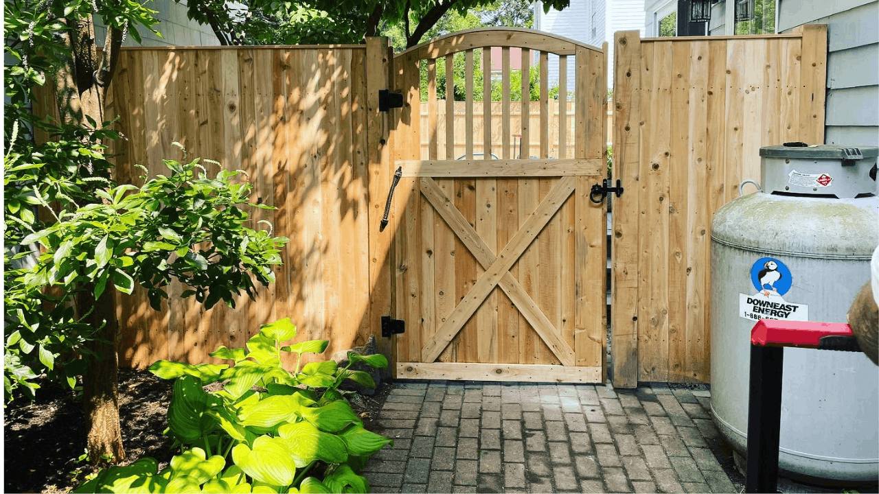 A wooden backyard gate with a latch, surrounded by a wood fence, with green plants and trees nearby, and a propane tank on the right side.