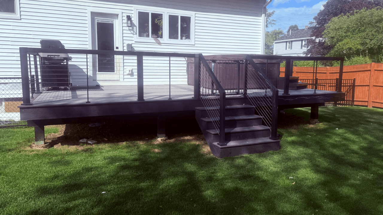 Backyard deck with stairs, black railing, white house in the background, grill, and grassy yard with trees.
