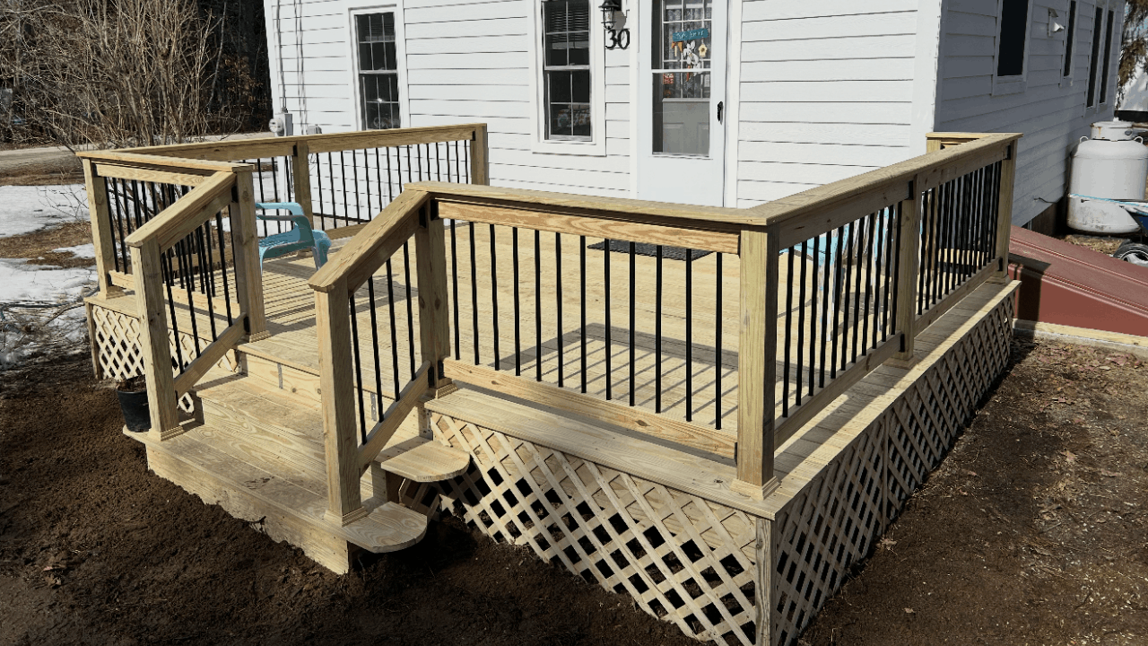 Newly built wooden deck with black metal railings attached to a white house, with stairs leading down to ground level, and a blue chair on the deck.