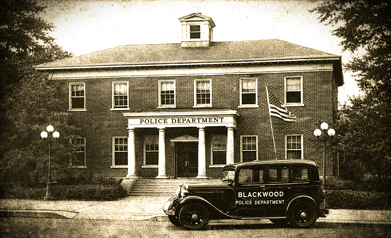 A vintage black police car labeled 'Blackwood Police Department' parked in front of a brick police station building with a sign that reads 'Police Department'. The building has a classical design with columns and an American flag on a flagpole. There are trees and street lamps around the building.