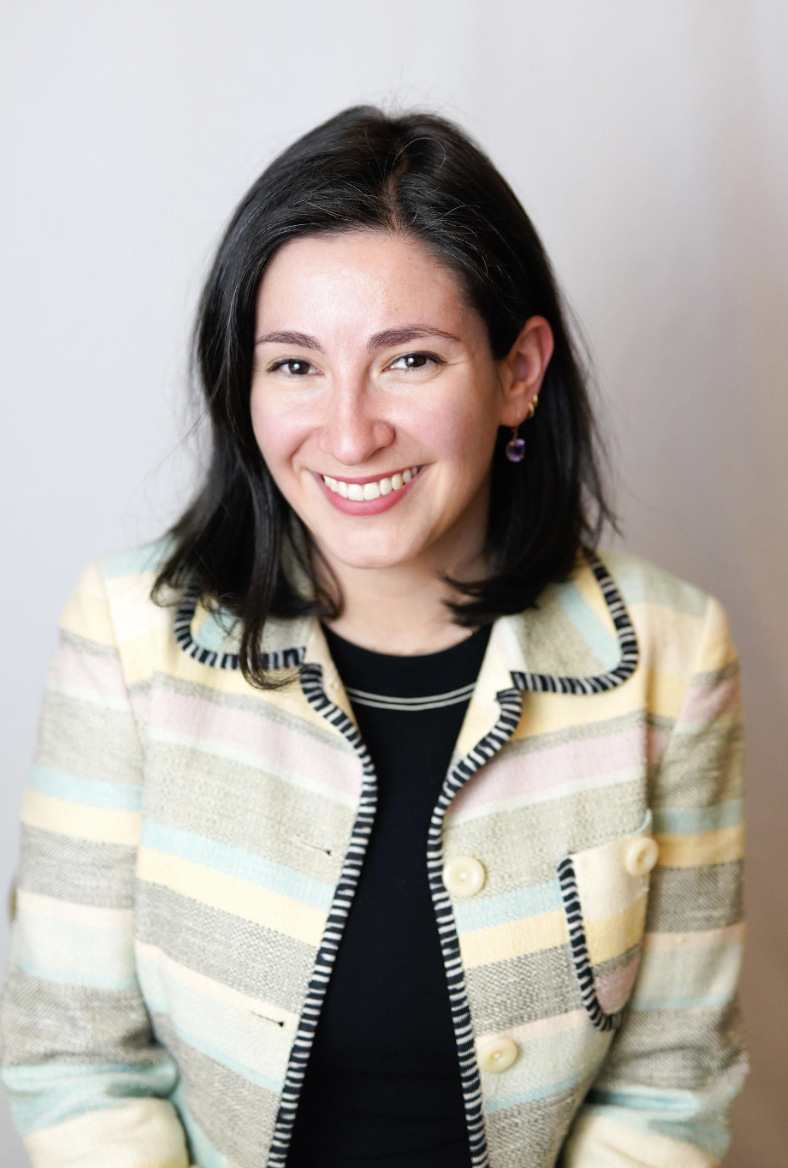 A woman (Dr. Ersilia Anghel) with black hair smiling, wearing a colorful striped blazer and a black top, standing in front of a plain background.