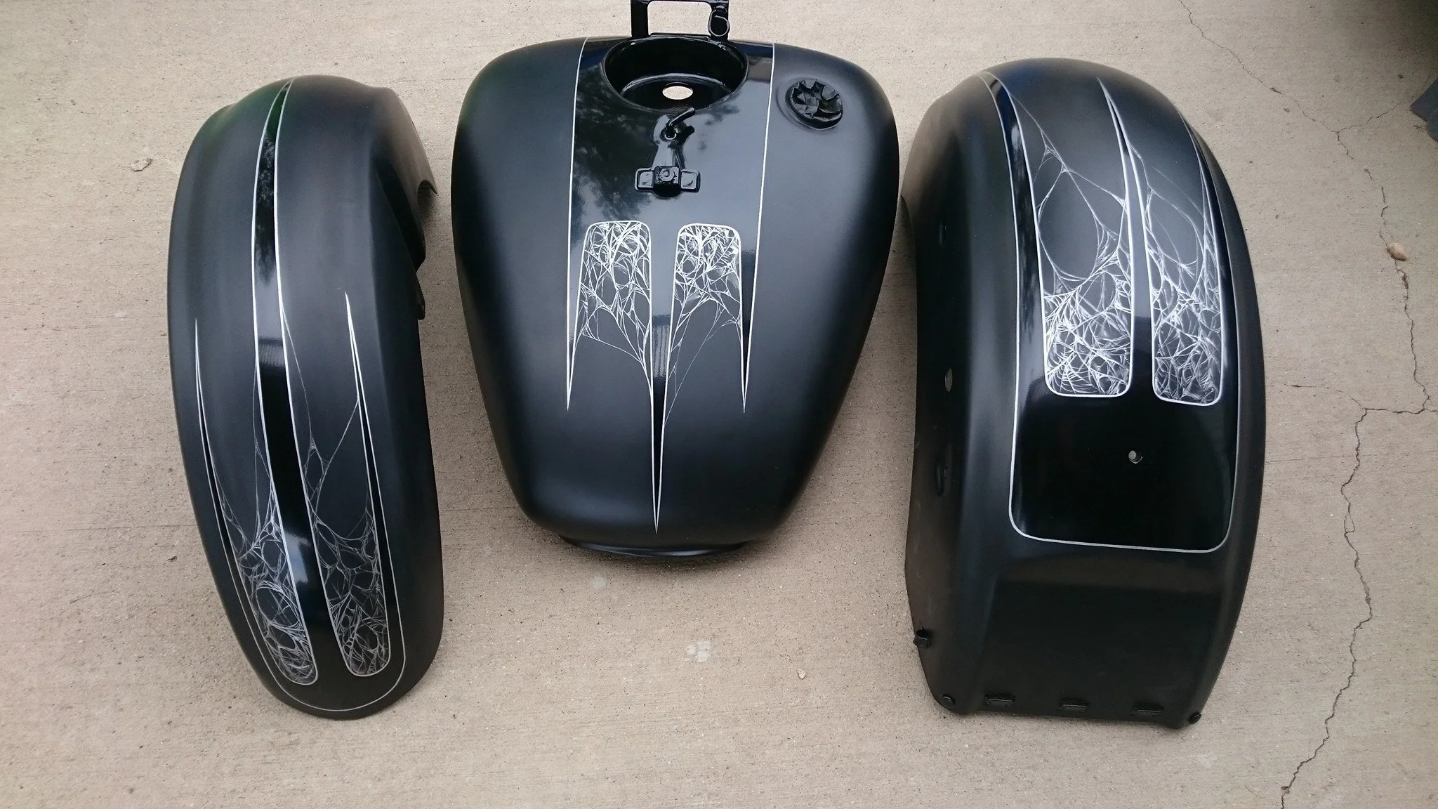 Three black motorcycle fuel tank and side panels with white spider web designs arranged on a concrete surface.
