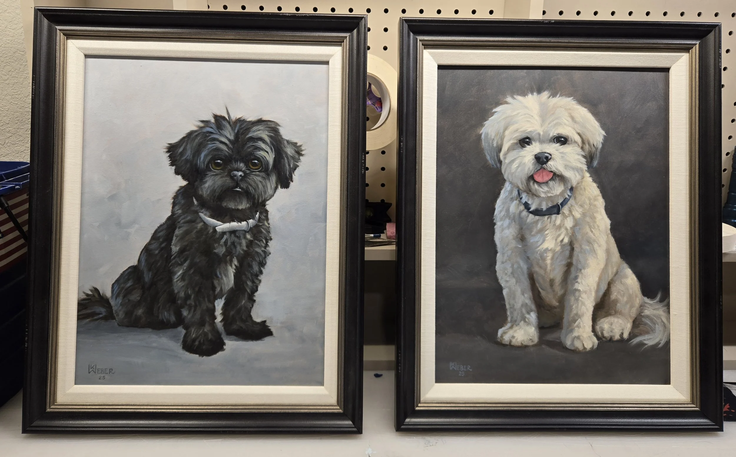 Two framed dog portrait paintings on a pegboard wall, one of a black dog and the other of a white dog, both sitting against a neutral background.