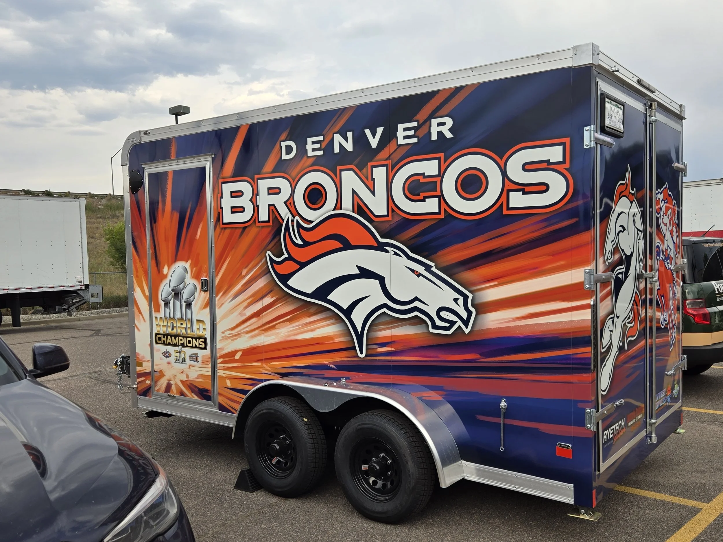 A Denver Broncos-themed trailer featuring the team's logo and colors, parked in a lot with a cloudy sky overhead.