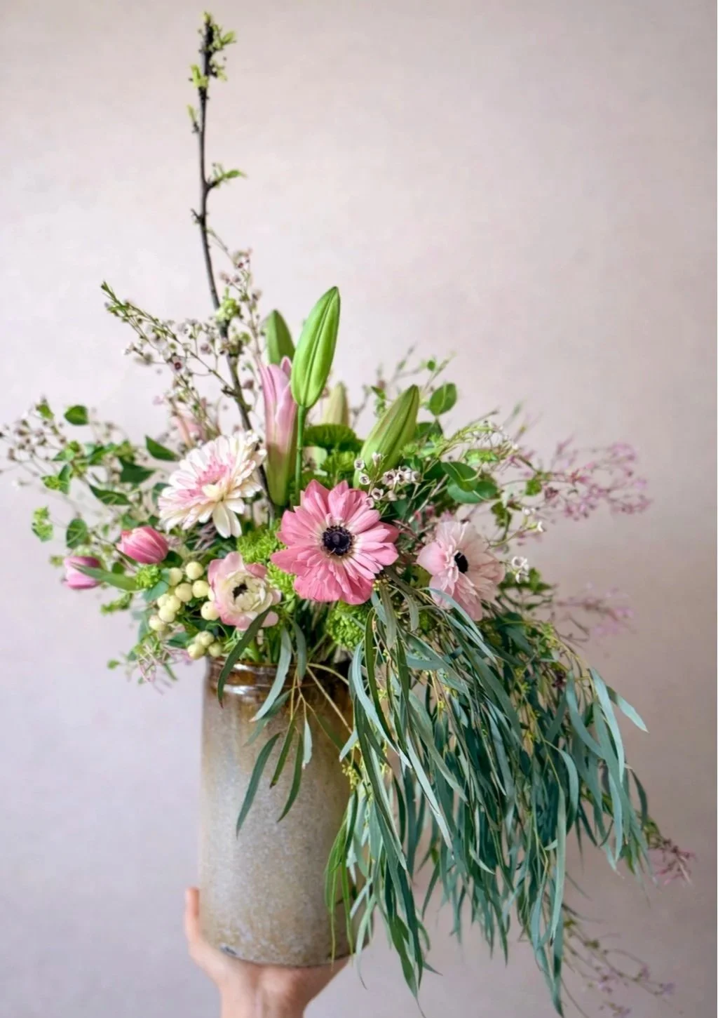 Hand holding a large beige vase filled with pink, white, and green flowers and greenery.