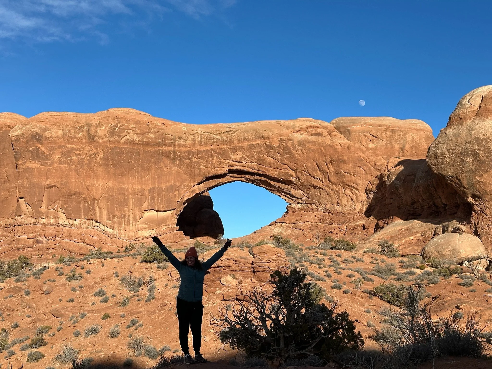 A person standing in front of a large natural rock arch formation in a desert landscape, with the moon visible in the clear blue sky.