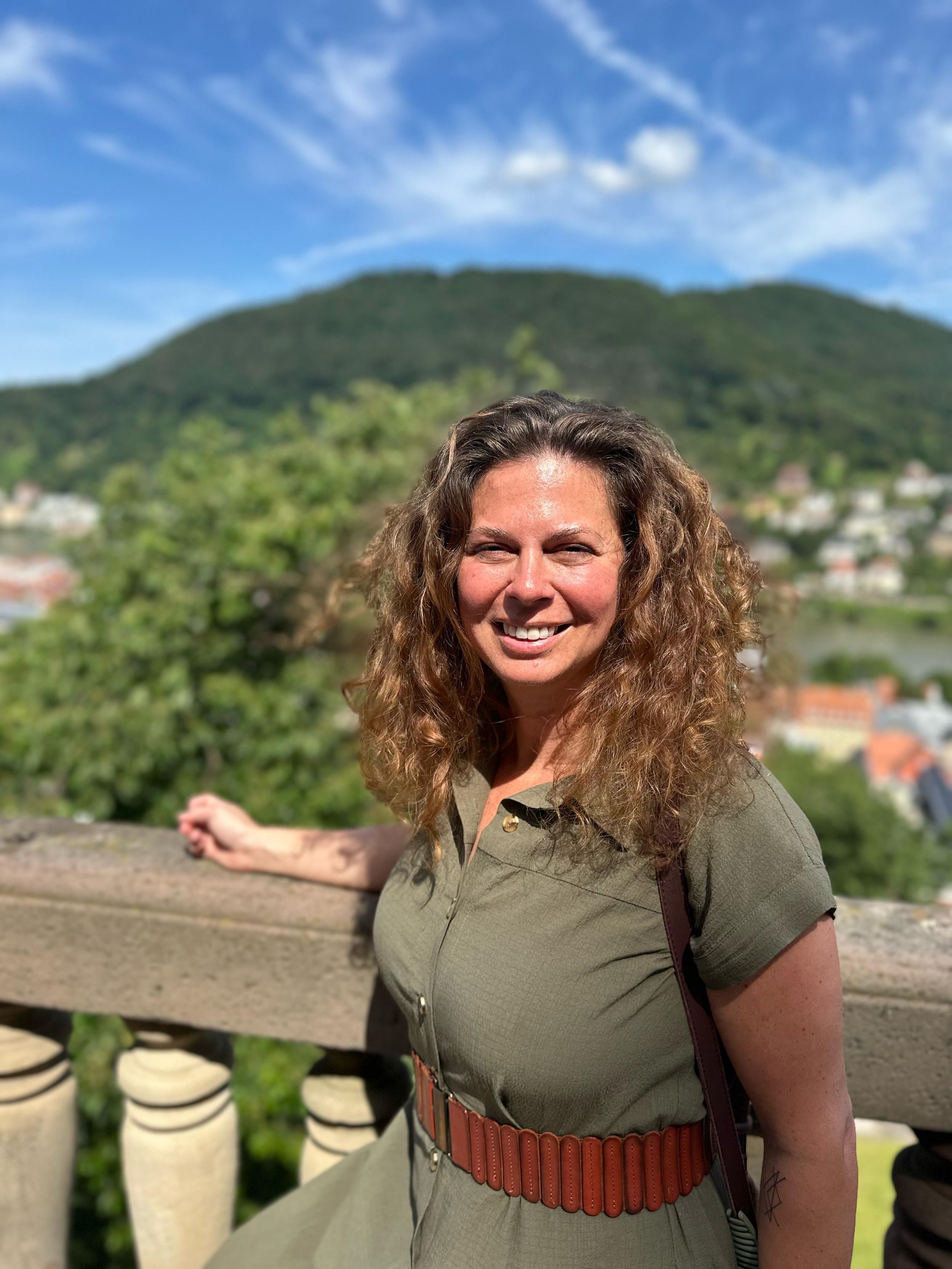 A woman with curly brown hair smiling outdoors, overlooking a green hill and distant town under a partly cloudy sky.