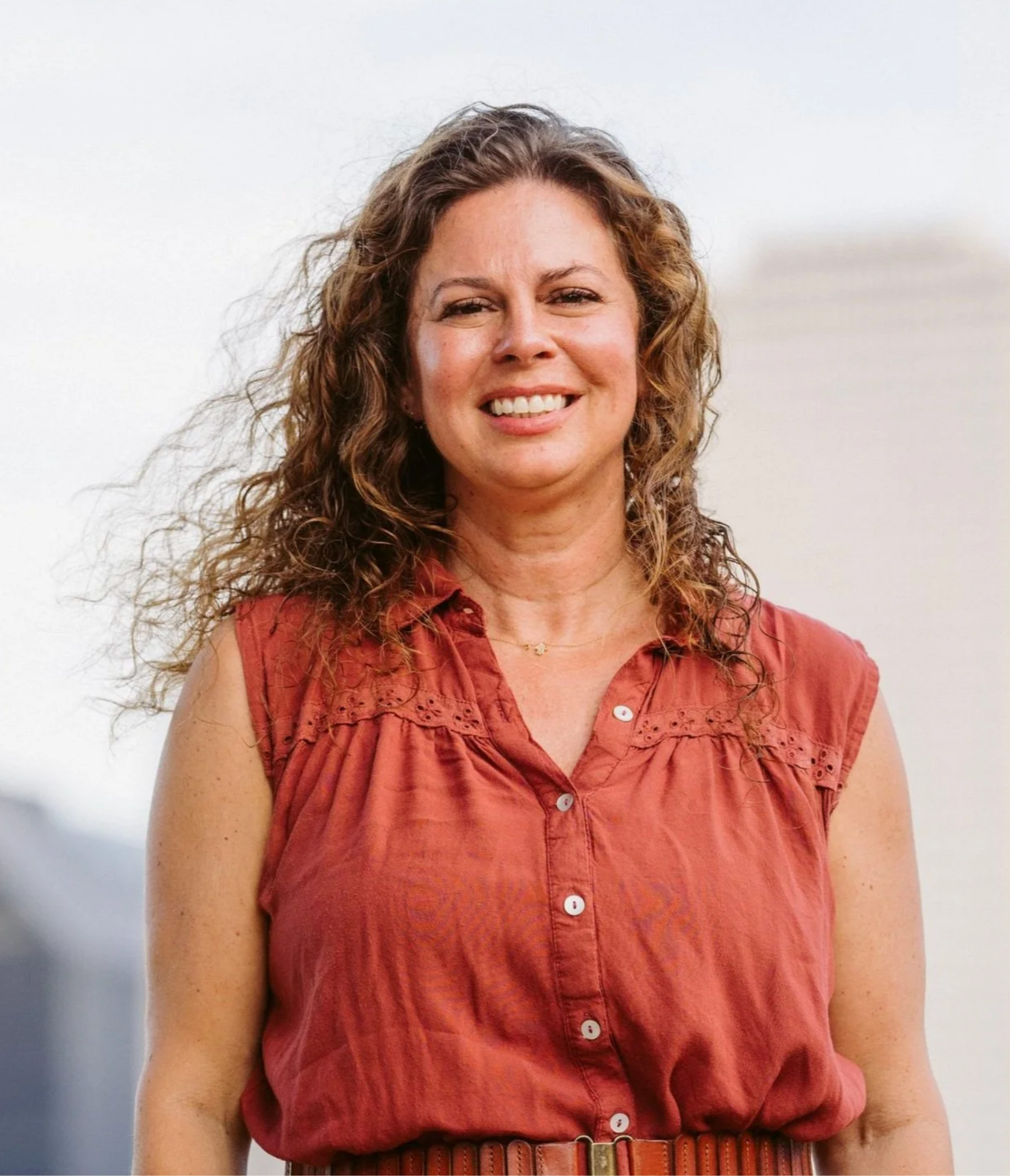 A smiling woman with curly brown hair wearing a sleeveless, rust-colored blouse, standing outdoors against a light sky background.