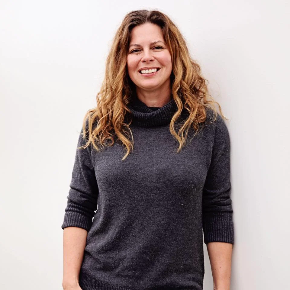 A woman with curly, reddish-brown hair smiling while standing against a white wall, wearing a dark gray turtleneck sweater.