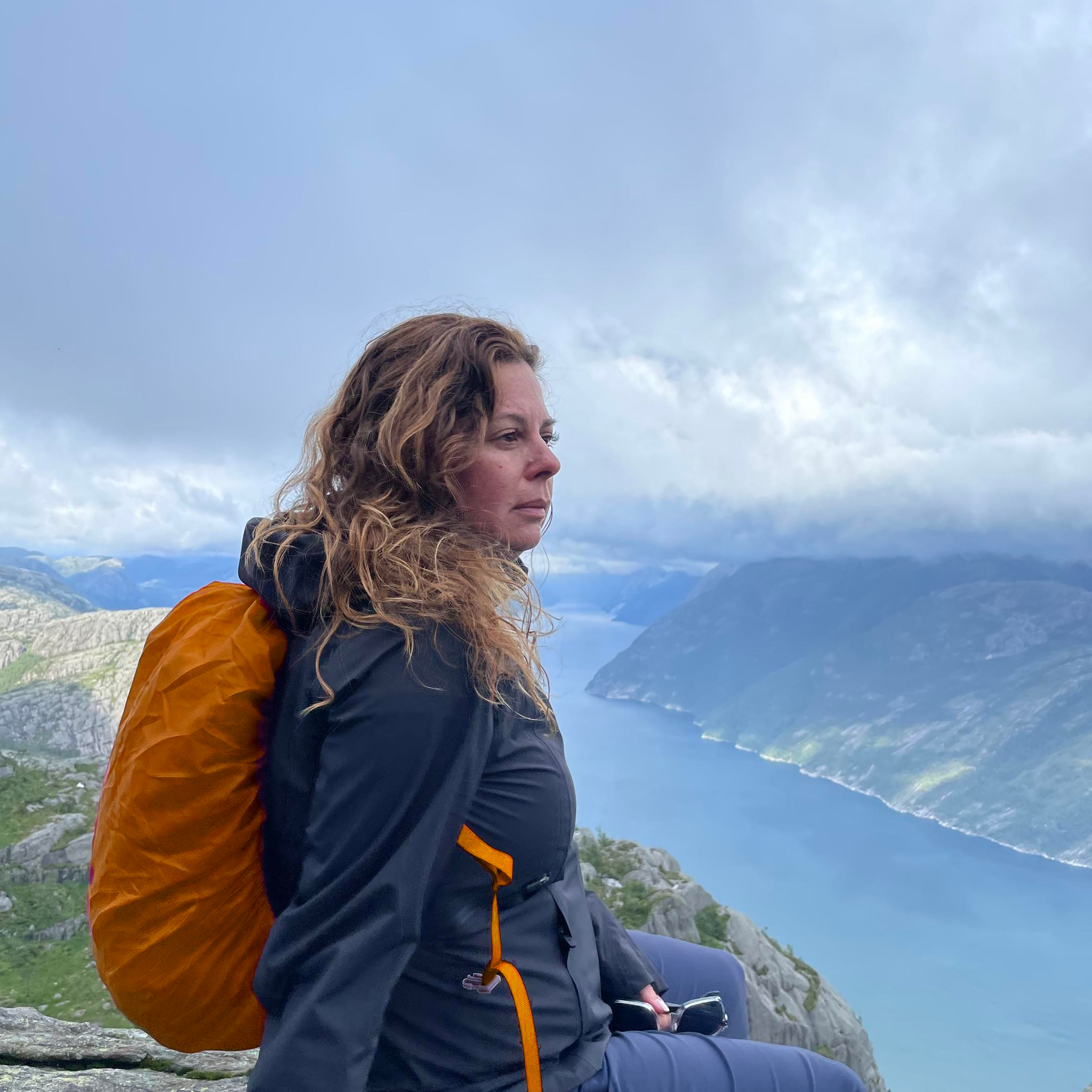 A woman with curly hair sitting on a rocky ledge overlooking a fjord with mountains and a cloudy sky in the background.