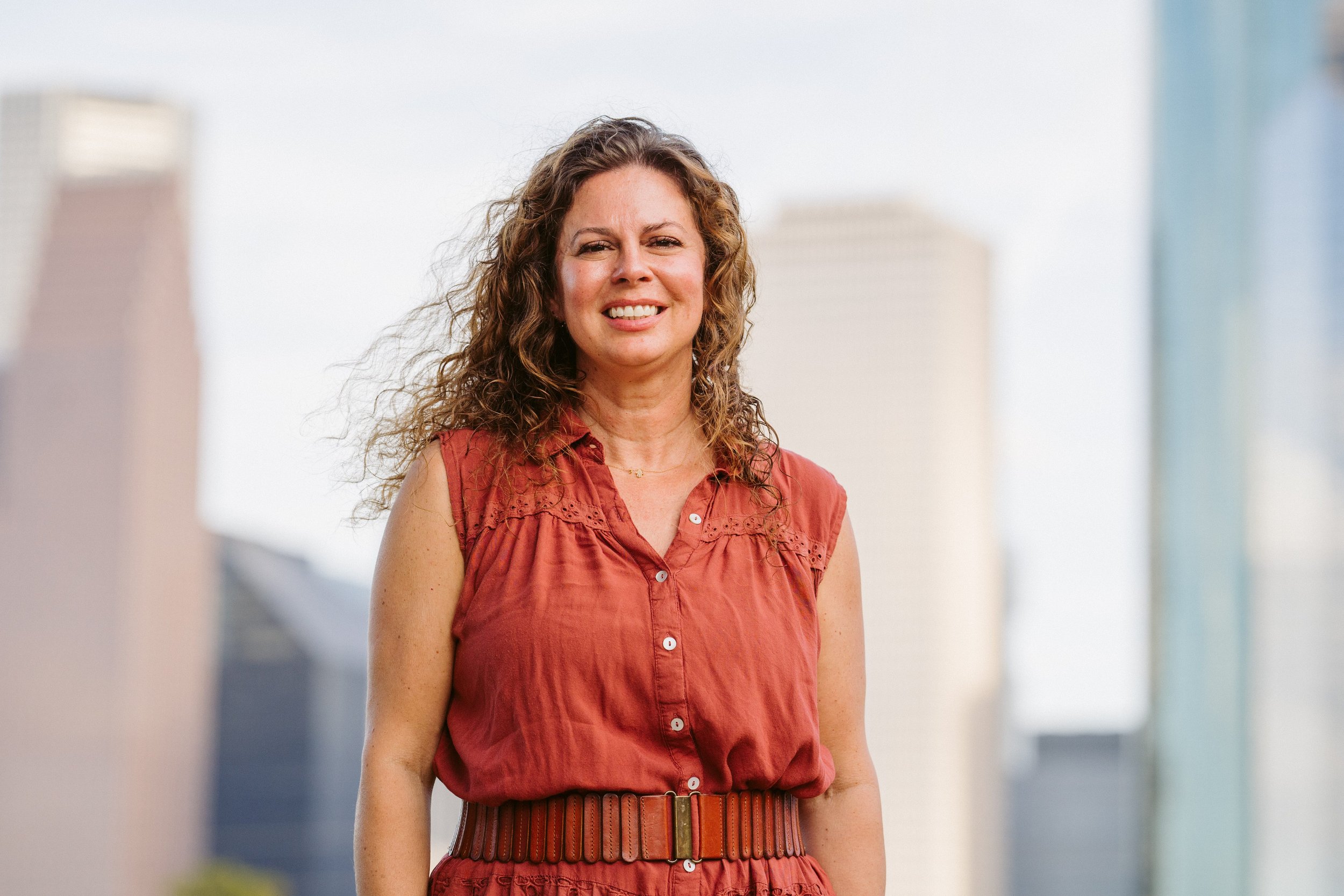A woman with curly brown hair smiling outdoors, wearing a rust-colored sleeveless button-up dress with a belt, against a cityscape background.