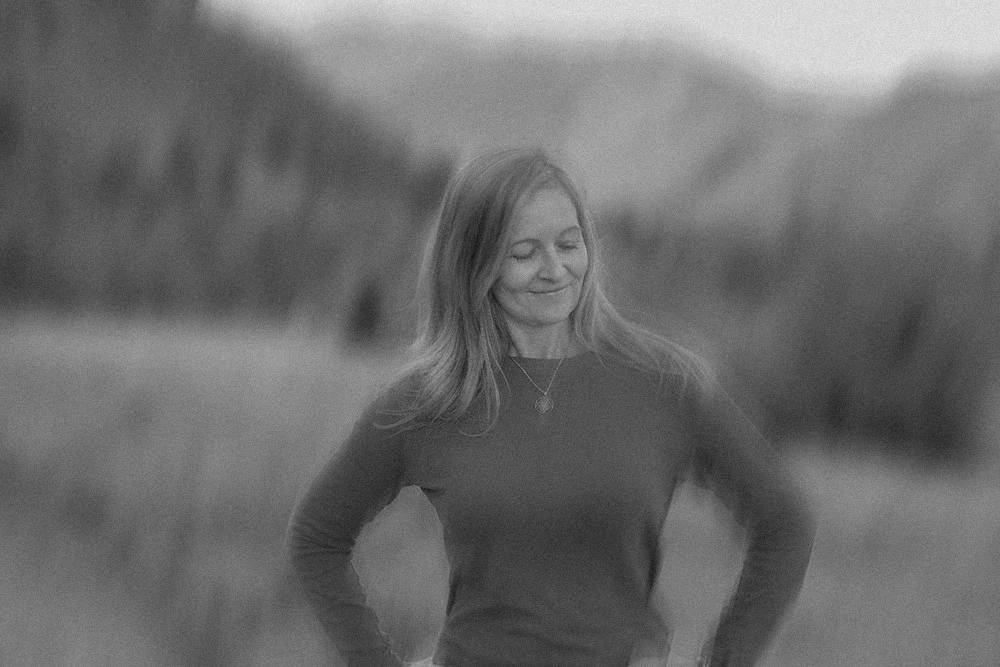A woman standing outdoors with her eyes closed and a slight smile, in a grassy field with blurred hills in the background.
