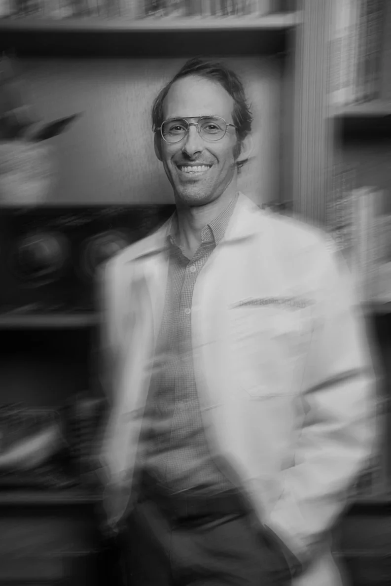 A smiling man wearing glasses and a light-colored jacket stands in front of shelves with books and items, with a motion blur effect.