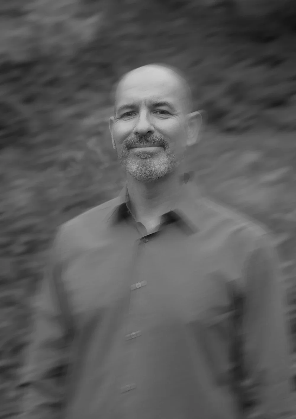 Black and white photo of a man with a beard, smiling, with blurred background that appears to be water, wearing a button-up shirt.