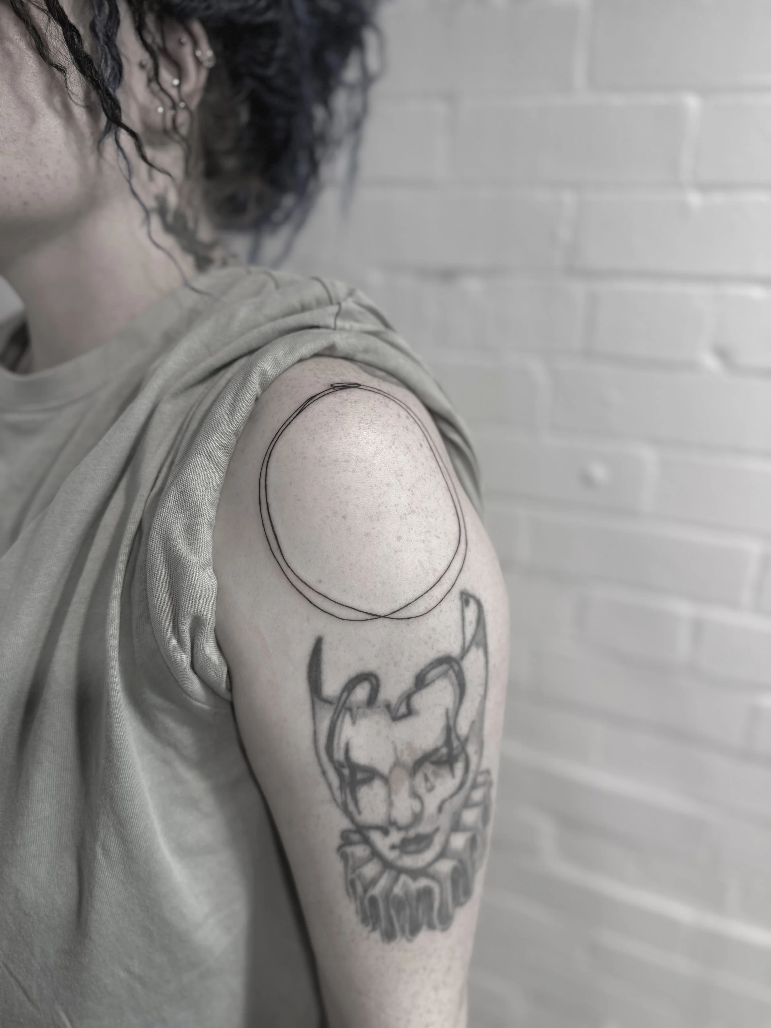 A person with tattoos on their shoulder and upper arm, including a woman’s face with devil horns and a crown, and a large circle outline near the shoulder, against a white brick wall.