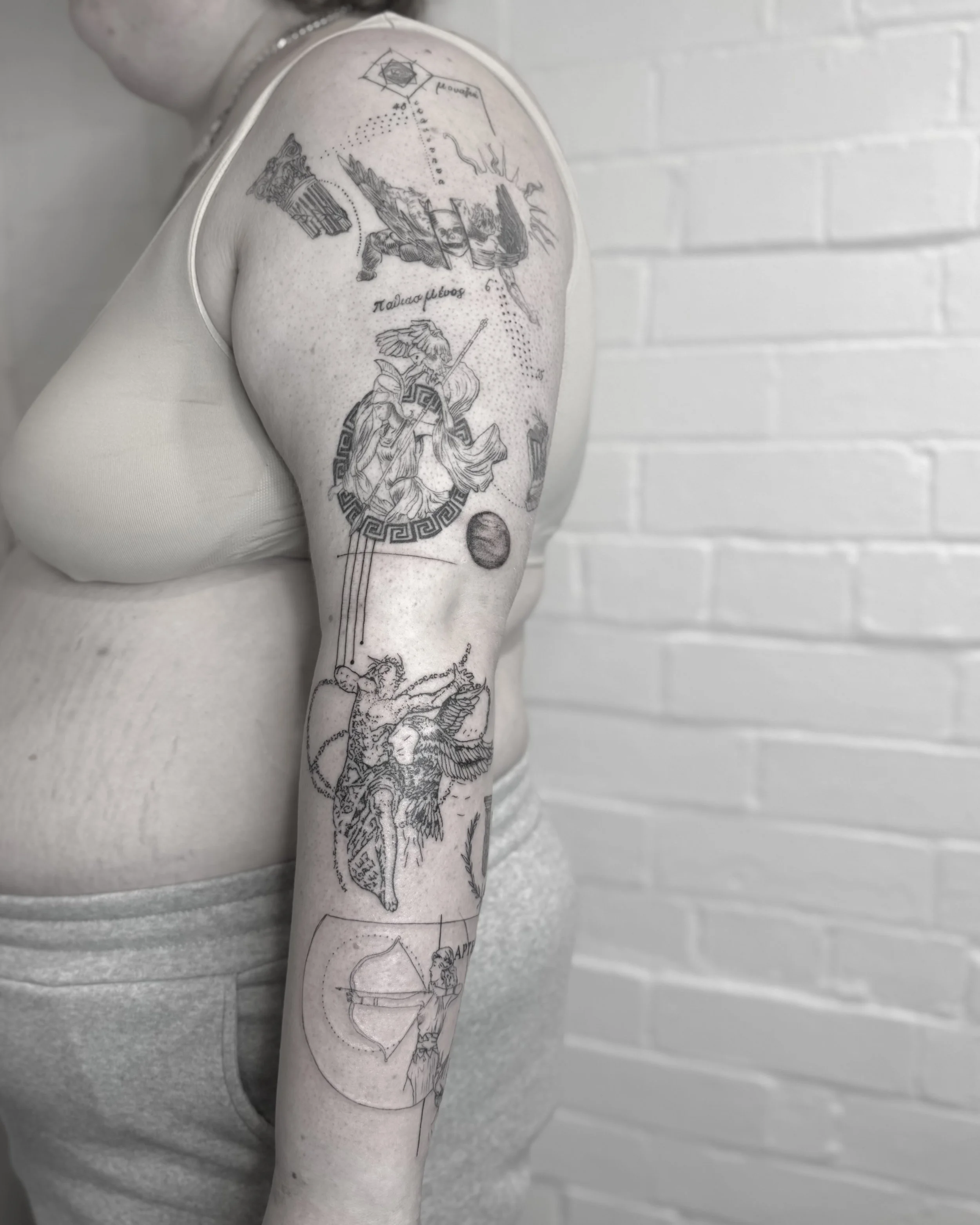 Black and white tattoo sleeve featuring various classical and mythological figures, geometric shapes, and Greek inscriptions on a person's arm.