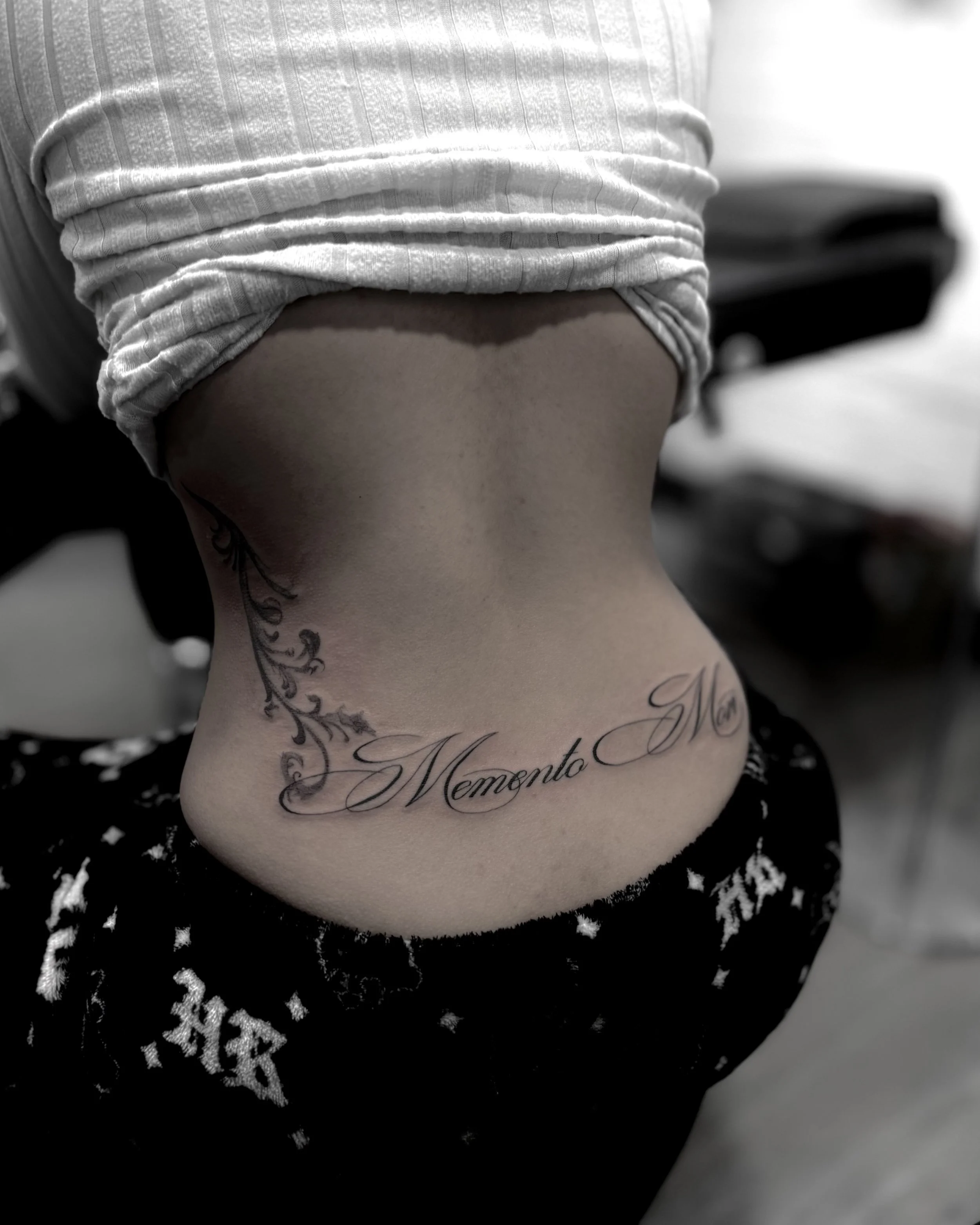 Close-up of a person's neck with a tattoo that reads 'Memento Mori' in cursive, surrounded by decorative floral designs, wearing a white shirt and dark patterned shorts.