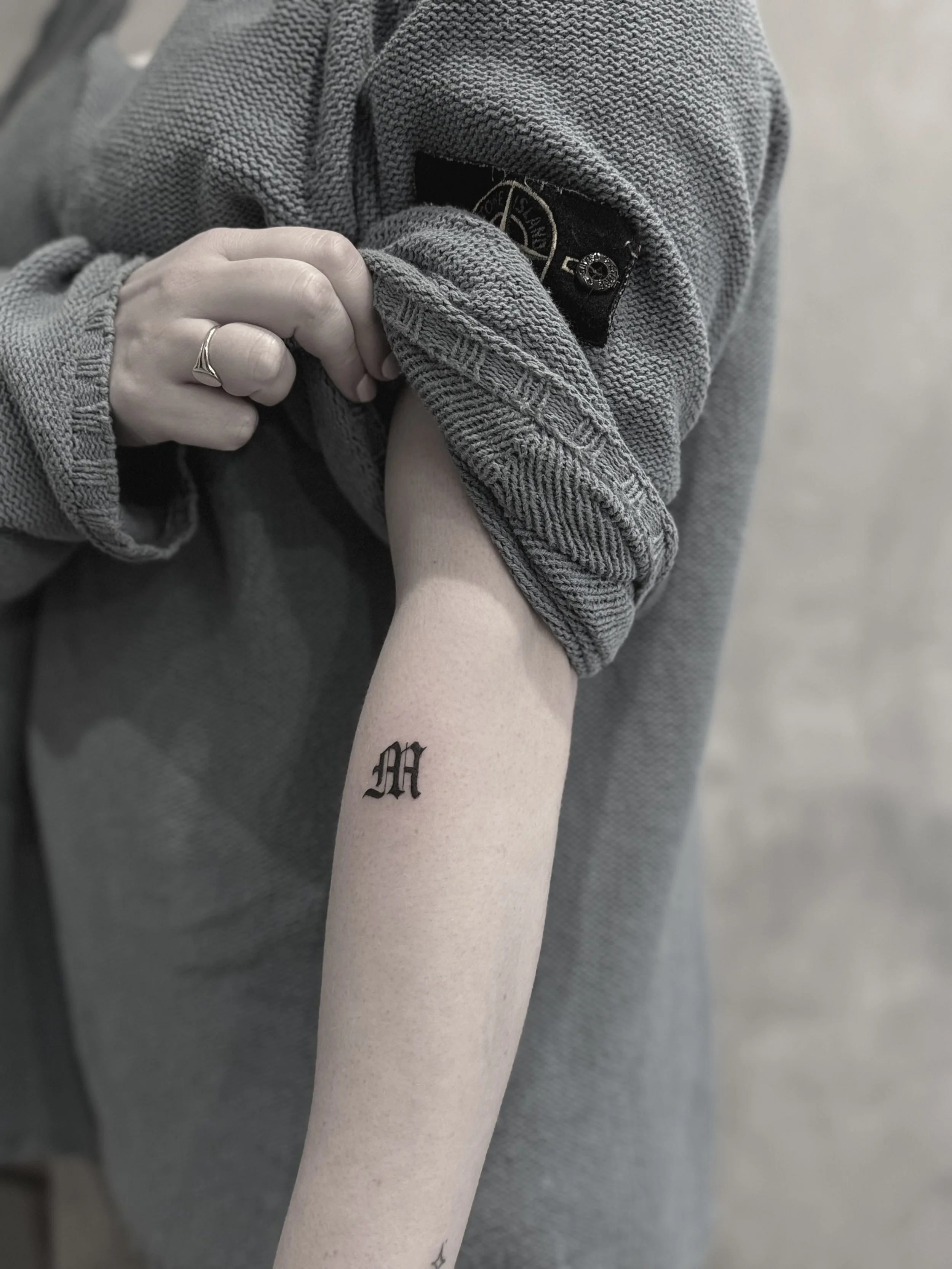 A person with a tattoo of an uppercase letter 'M' on their forearm, wearing a gray jacket with rolled-up sleeves, reveals the tattoo by lifting the sleeve.
