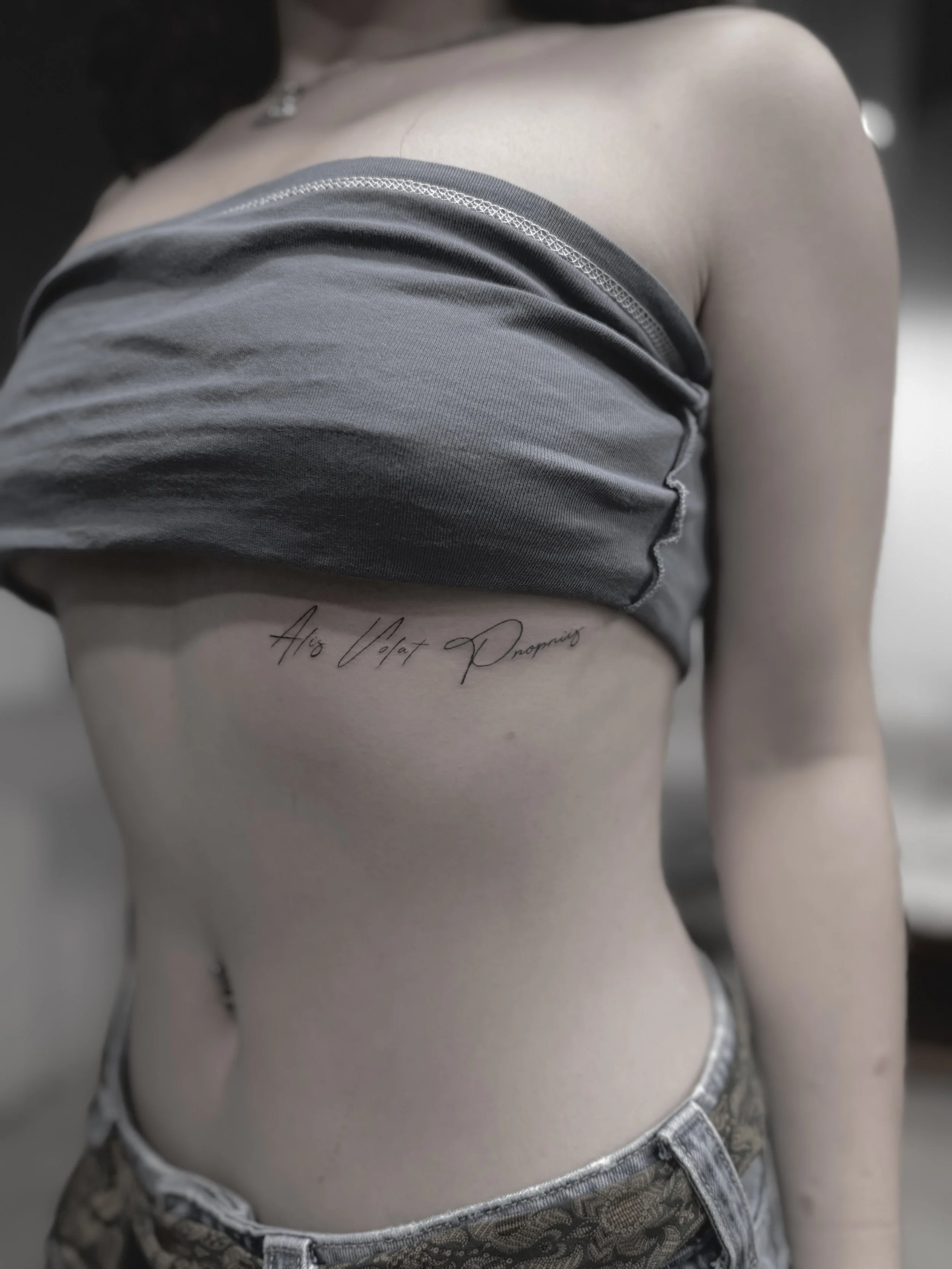 Partial view of a woman's torso with a tattoo in cursive reading 'Alo Volat Dropping', wearing a gray crop top and patterned jeans.