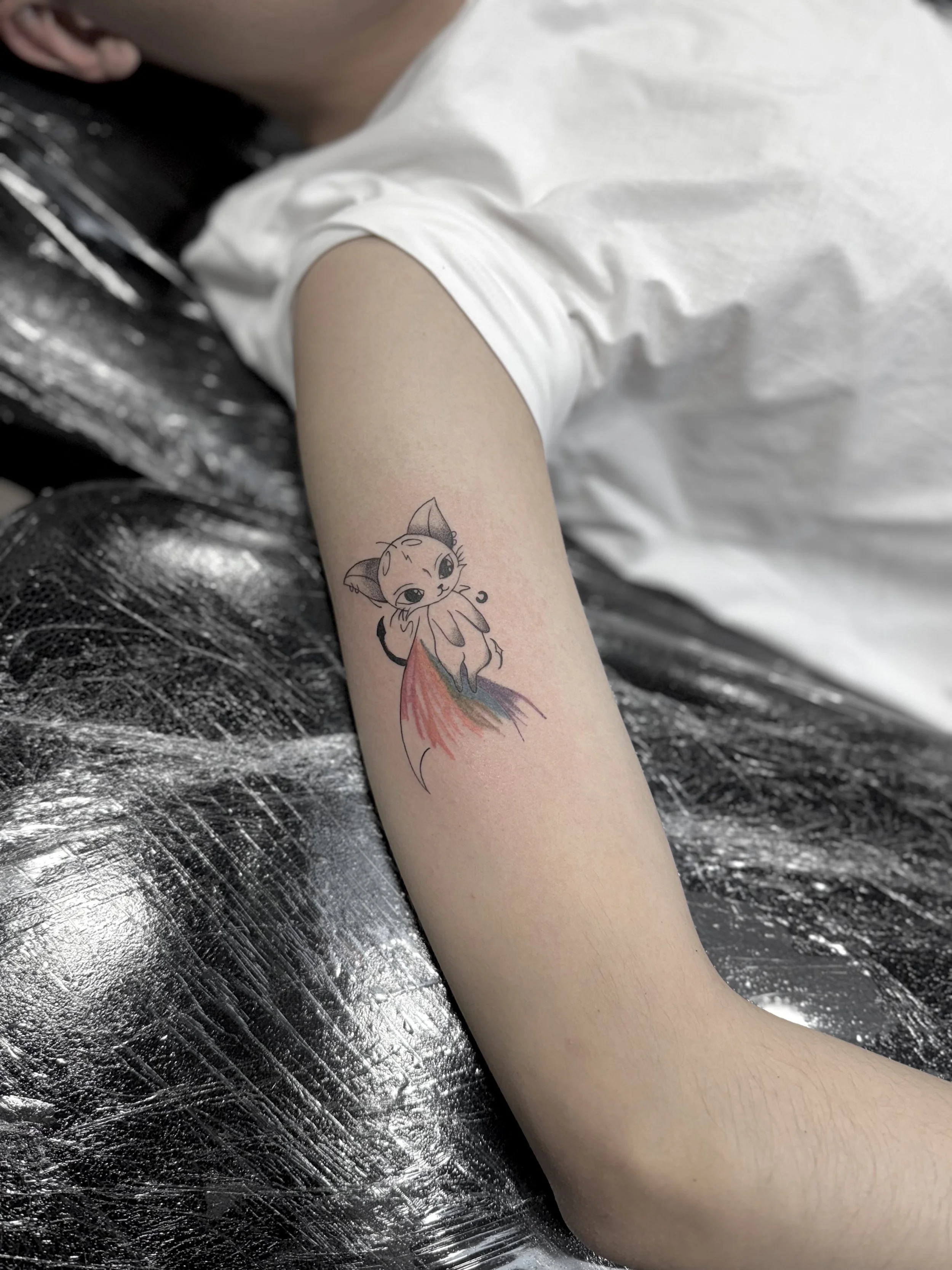 Tattoo of a cute cartoon kitten with a colorful tail on a person's arm.