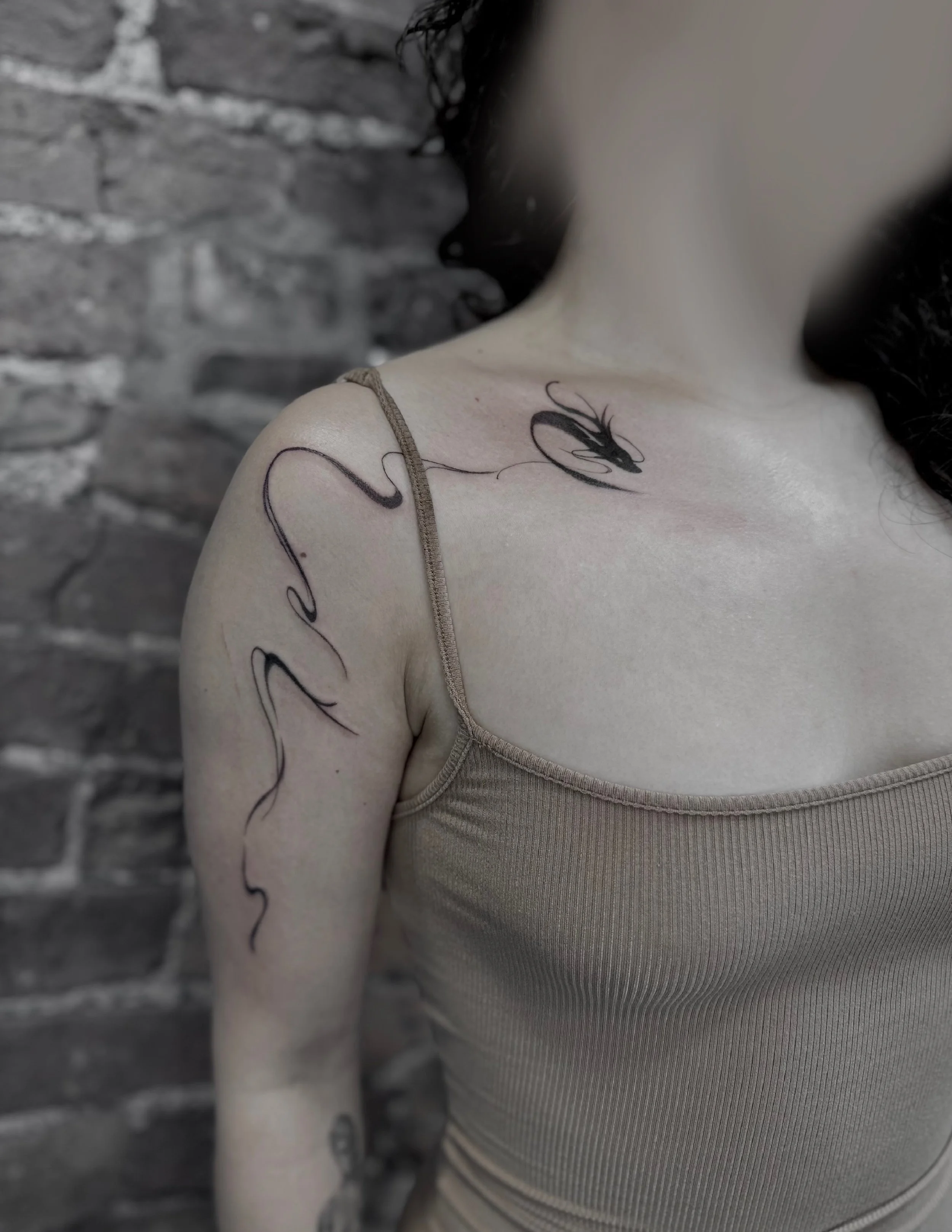 A woman with black hair wearing a beige tank top, showing a tattoo of a swan on her chest and abstract line art on her shoulder, against a brick wall background.