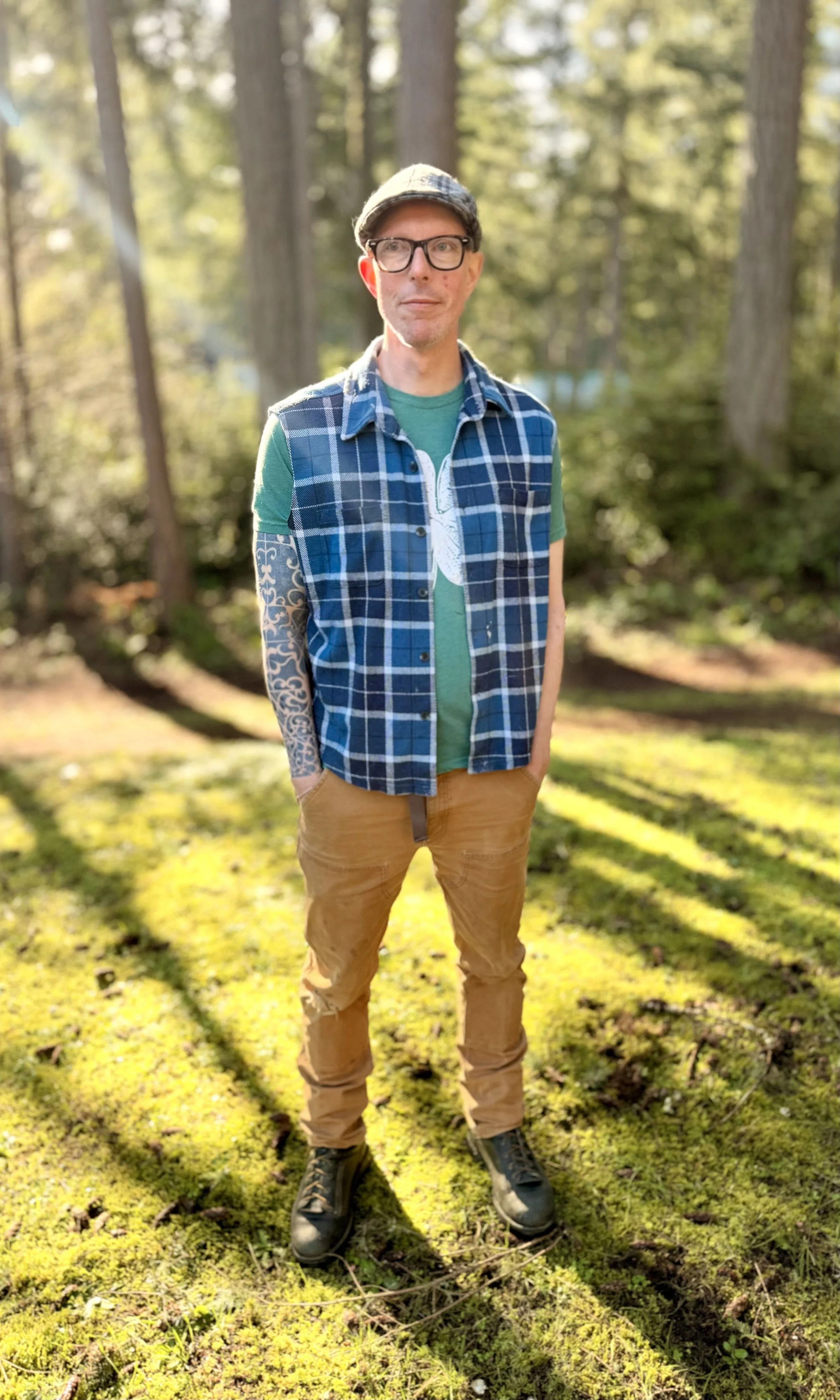 A man standing outdoors in a forest with sunlight filtering through trees, wearing a plaid sleeveless shirt over a green t-shirt, tan pants, black boots, glasses, and a cap.