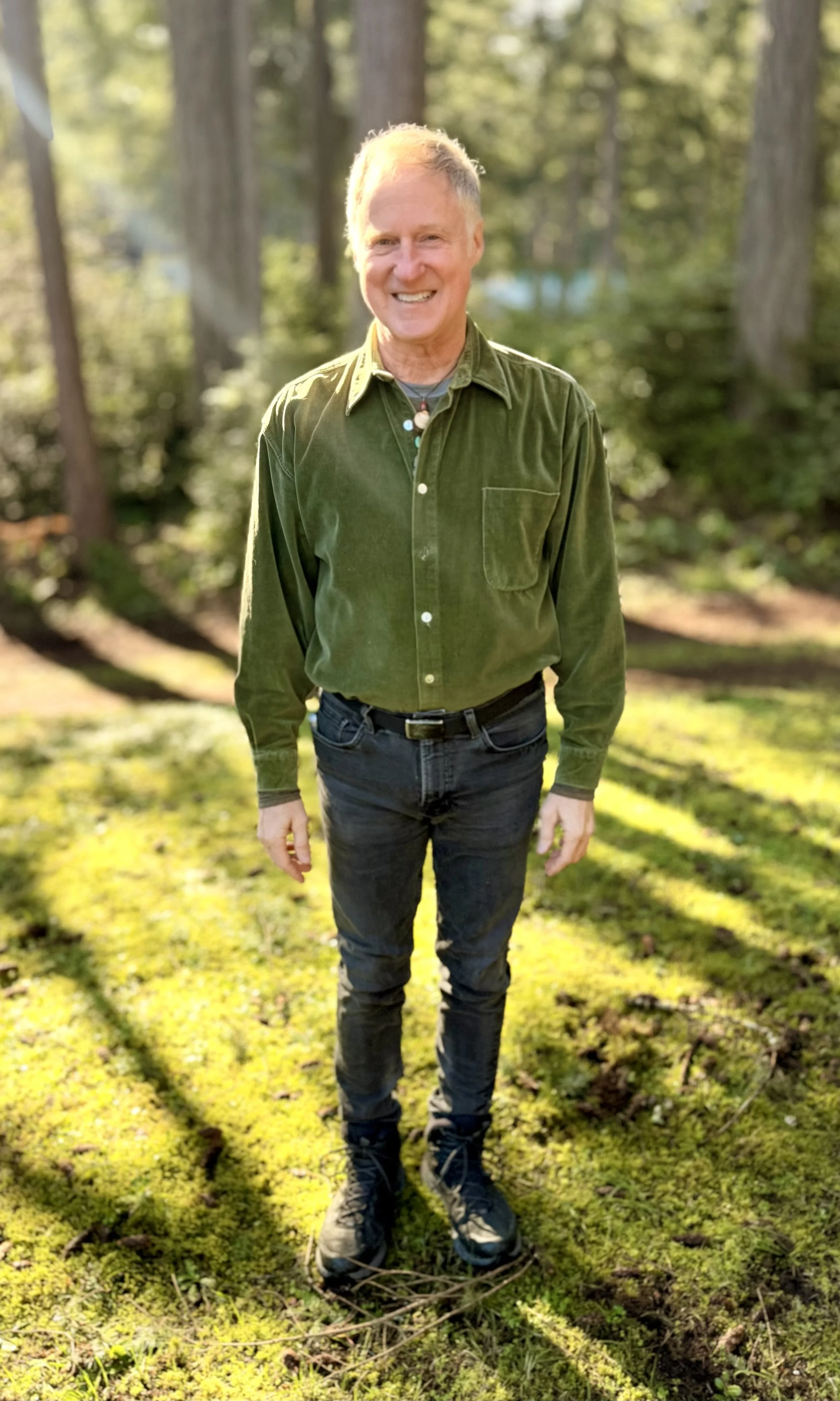 A smiling man with short gray hair standing outdoors on a mossy ground with trees in the background.